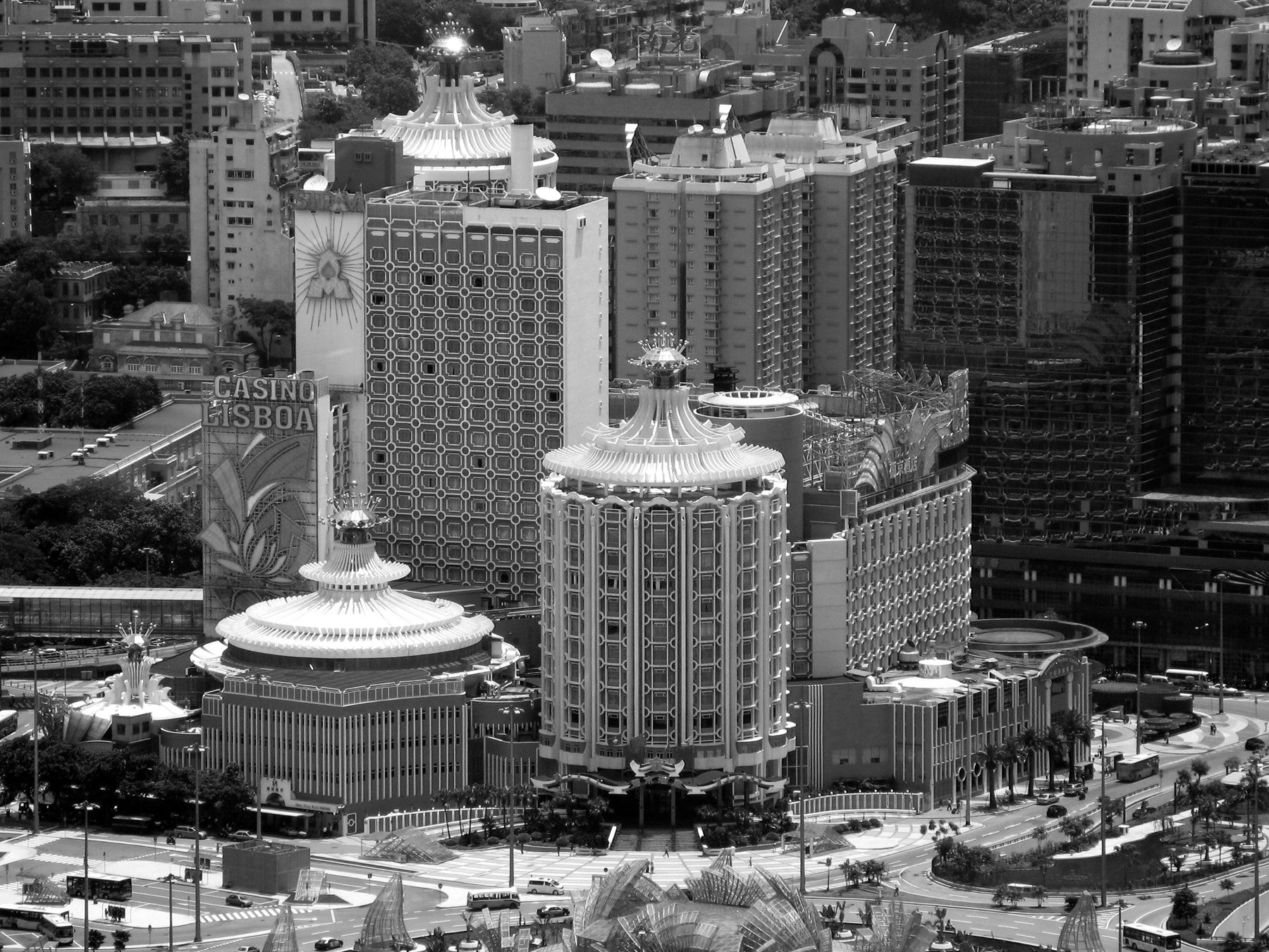 Looking down on a cluster of tall hotels and a domed, crown-topped casino building at a busy road junction in a dense city.