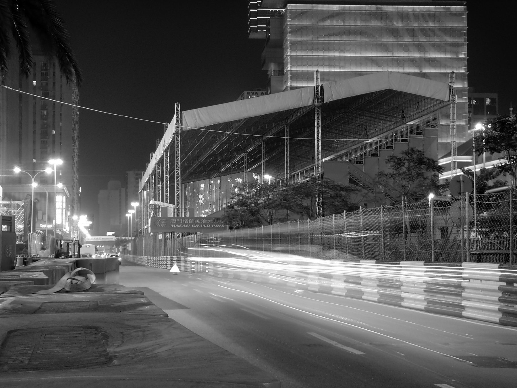 At night, the light trails of passing cars streak along a street circuit lined with barriers, beneath a covered grandstand and the lit towers of the city.