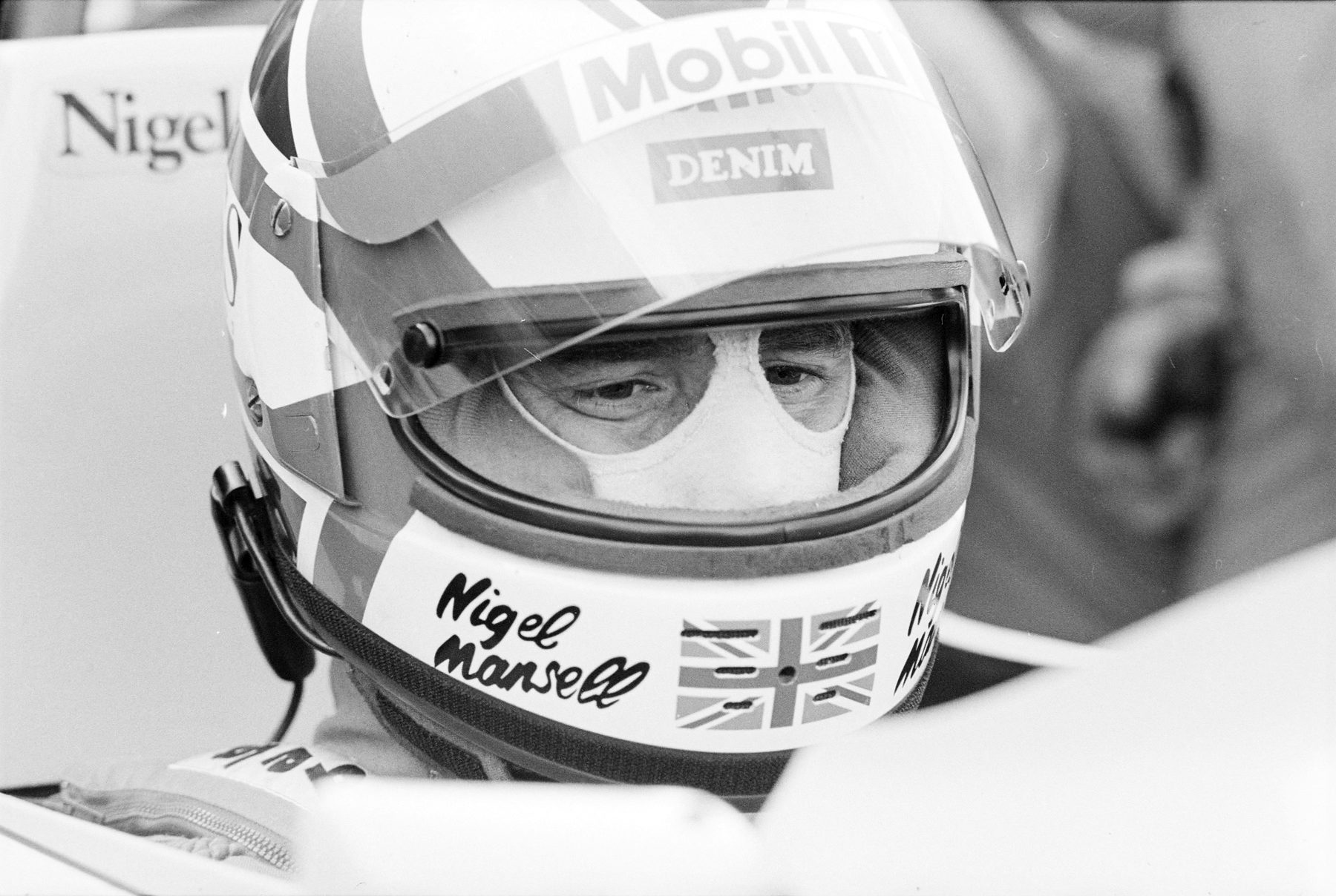 Close-up of a driver in a full-face helmet with the visor up, his name and a Union flag on the chin guard, sitting in his car in the pit lane.