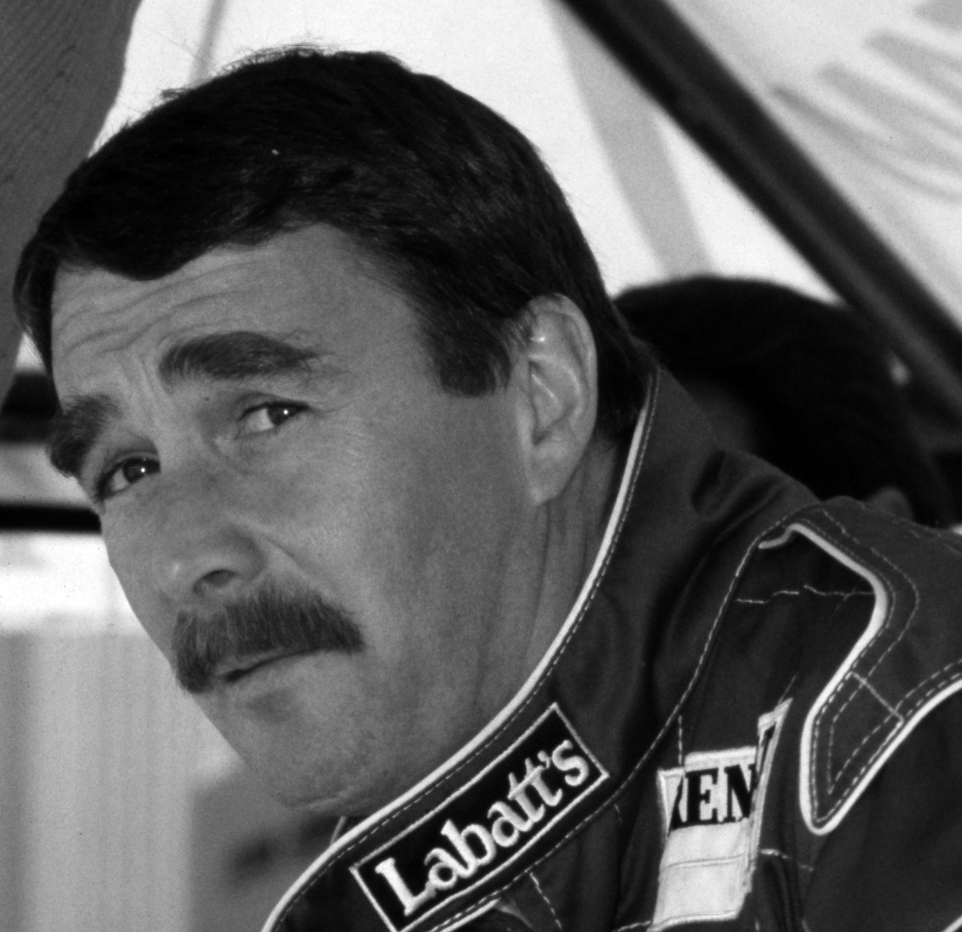 A dark-haired driver with a thick moustache, in dark team overalls with sponsor patches, ‘Labatt’s’ on the chest, looks towards the camera with a wary half-smile.