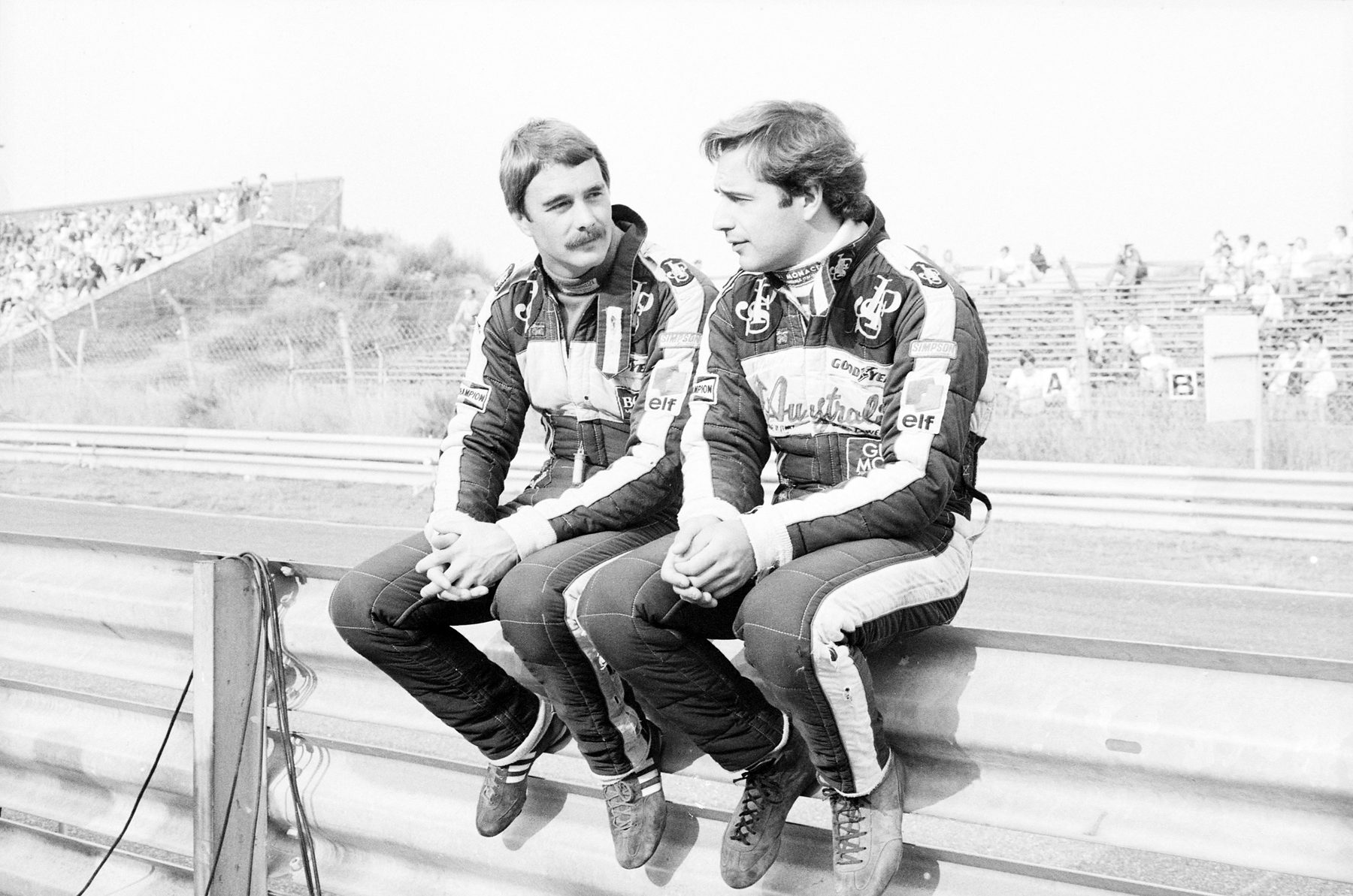 Dos jóvenes pilotos con trajes iguales de su equipo, sentados uno junto a otro sobre una barrera junto a la pista, uno de ellos con bigote, conversan; detrás, un talud de espectadores.