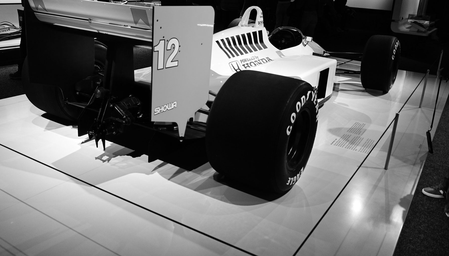 Seen from behind in a darkened hall, a pale Formula One car with a tall rear wing numbered 12 stands spotlit on a white plinth.