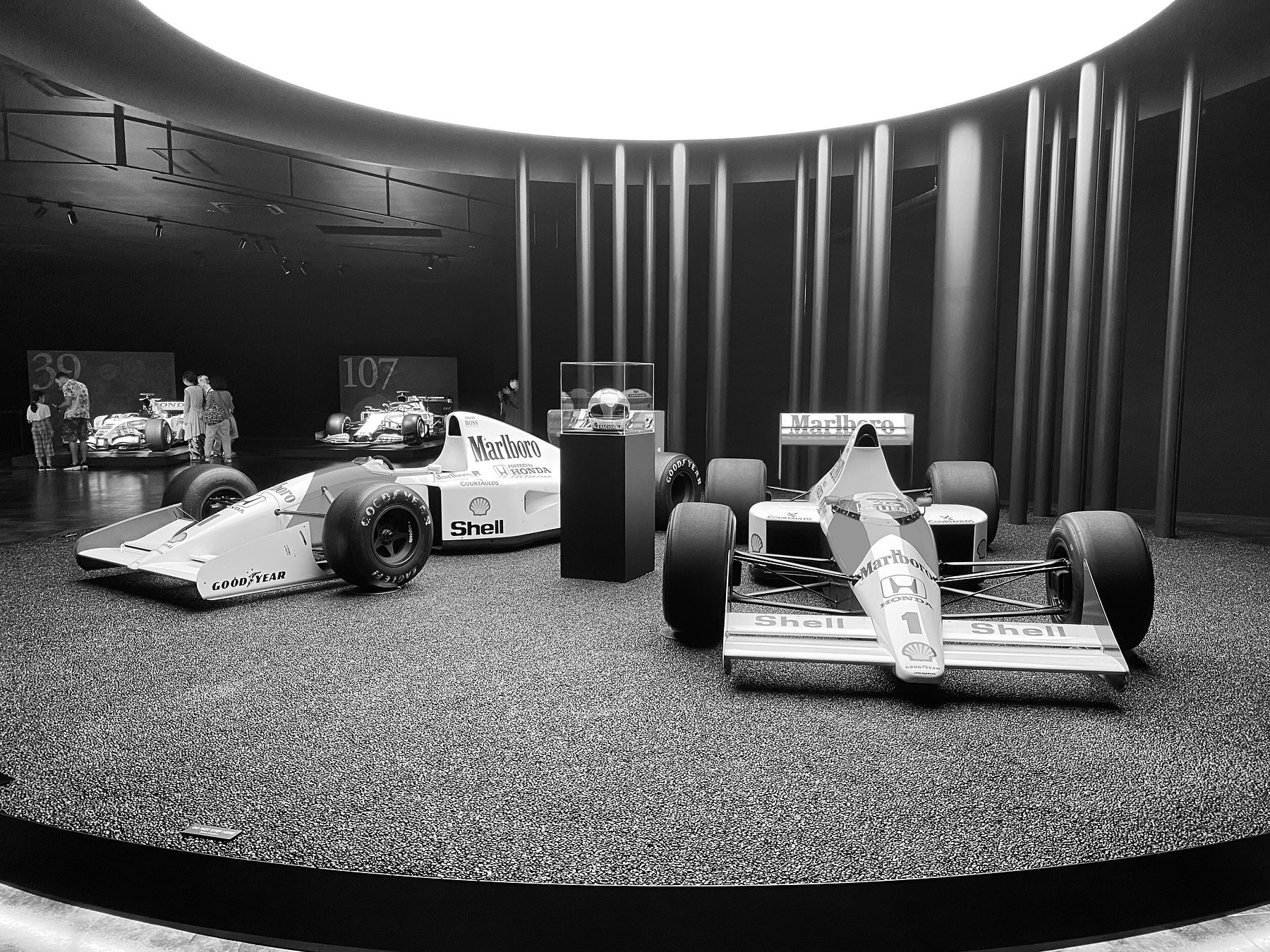 In a dark, circular hall under a ring of light, two pale Formula One cars stand on a gravel floor, one in profile and one head-on, with a helmet in a glass case on a plinth between them.