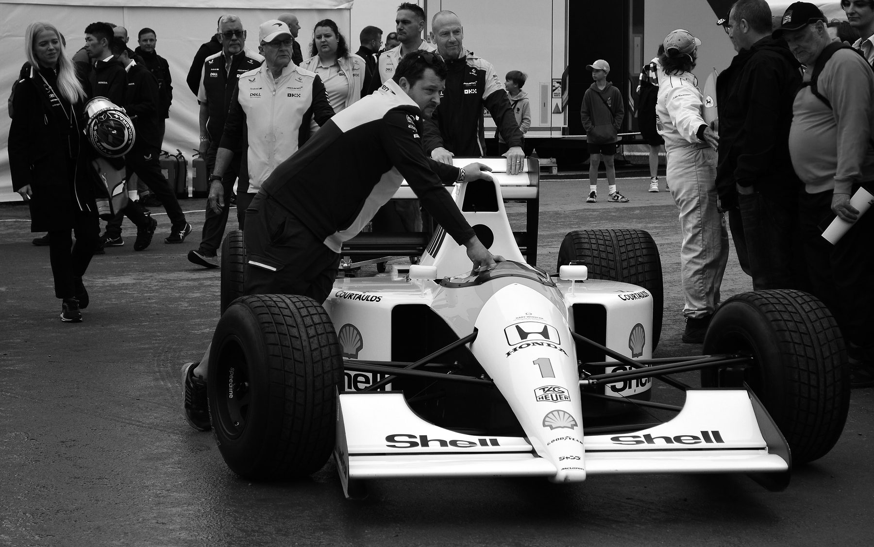 A mechanic in team kit leans on the roll hoop to push a pale Formula One car numbered 1 through a crowded paddock on a grey day, onlookers all around.