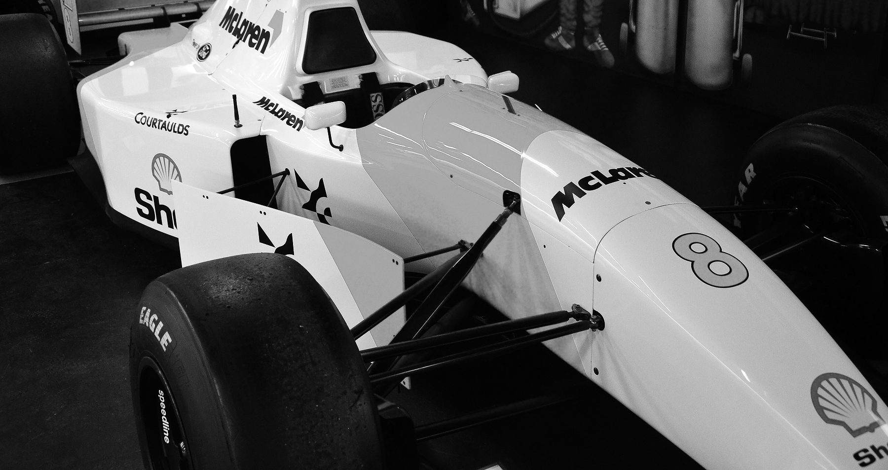 A pale Formula One car numbered 8, lettered ‘McLaren’ and ‘Shell’, seen from above and to one side in a dim museum hall, its cockpit and raised airbox towards the back of the frame.