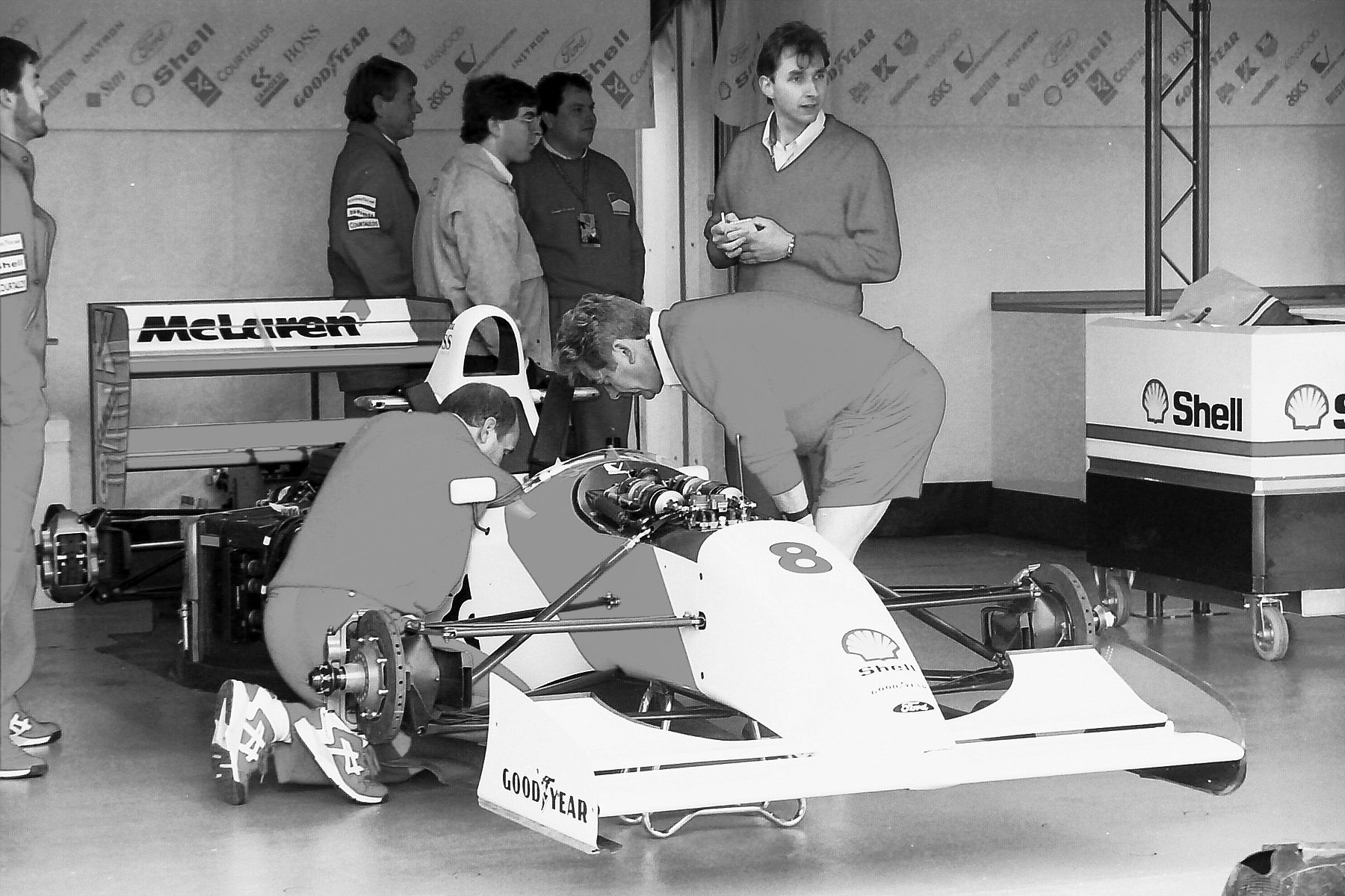In a garage, two mechanics crouch and lean over a pale Formula One car numbered 8, its engine cover off and its front wheels removed, while five more people stand and watch.