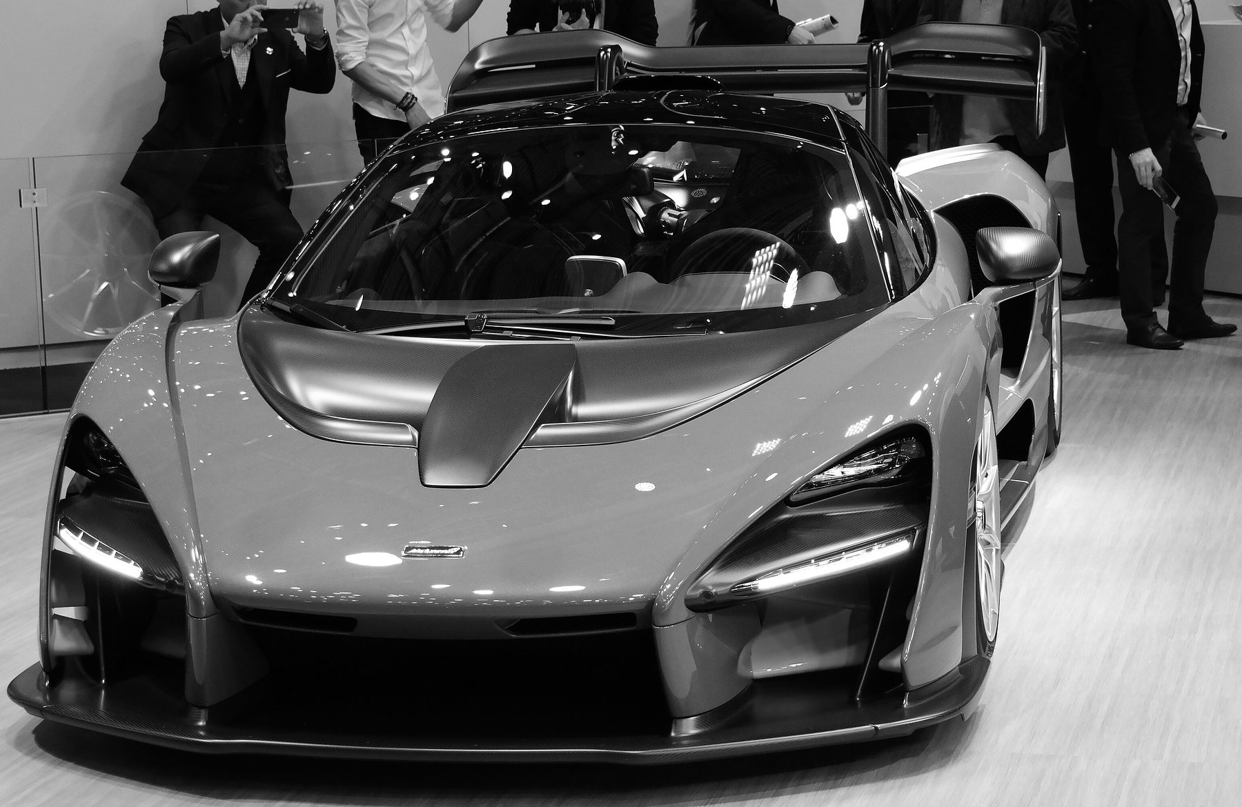 A low, wedge-shaped supercar in a mid-grey finish, with huge air intakes and a tall rear wing, stands on a show stand, visitors with phones crowding behind it.