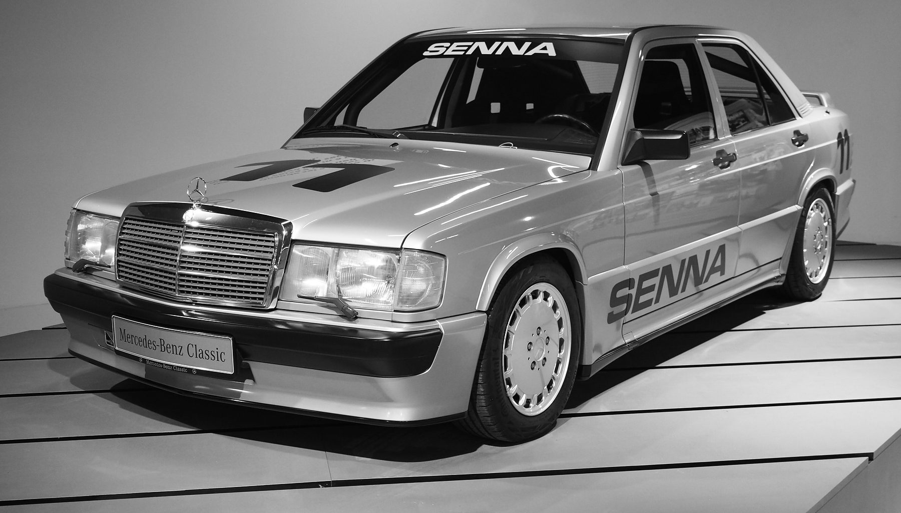 A pale 1980s four-door saloon with ‘Senna’ lettered on the windscreen and along the doors and a race number on the bonnet, displayed on a pale plinth.