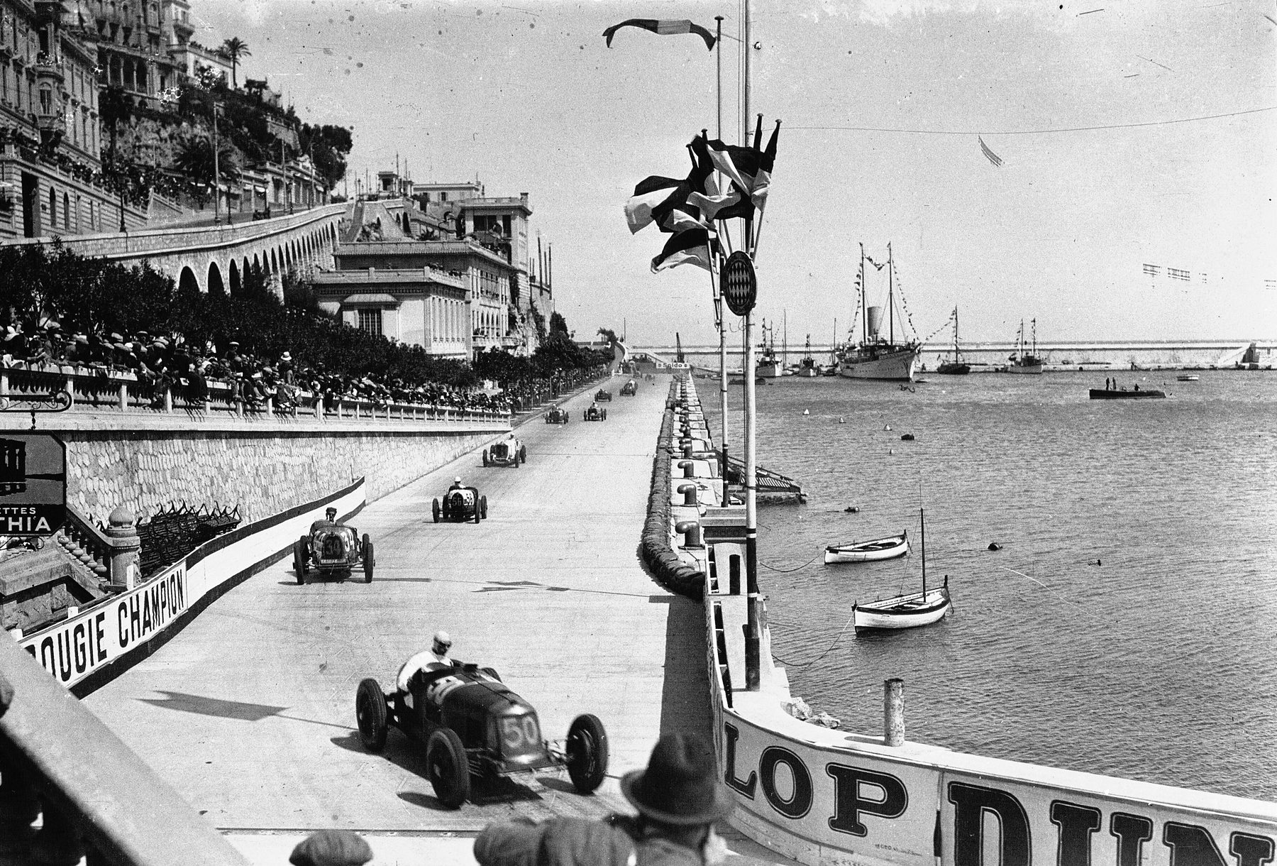 Racing cars of the early 1930s string out along a harbour-front road lined with advertising boards and crowds, yachts moored in the harbour at right.