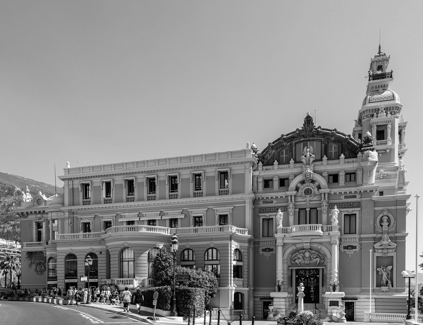 An ornate building with a dark-domed roof and a tall corner tower stands above a curving road; a line of visitors walks along the pavement past its hedges.