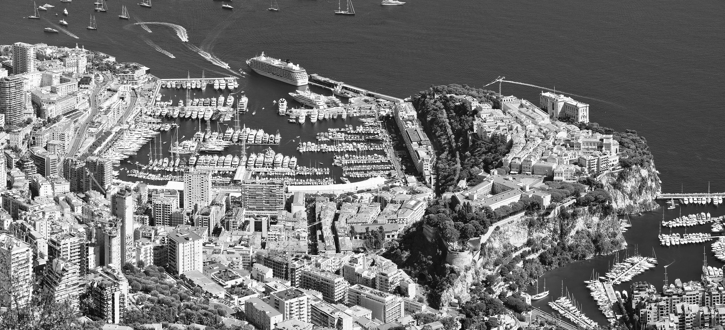 A panorama from high above: a harbour crowded with yachts, the old town on its rocky headland, and dense apartment blocks rising from the sea up the hillside.