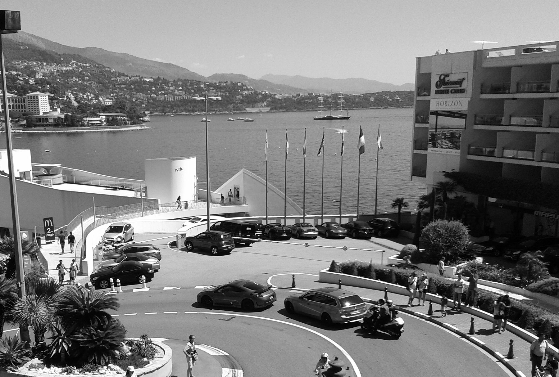 Looking down on a tight hairpin bend with striped kerbs at the foot of hotel buildings, cars and pedestrians crossing it, the sea and the coast beyond.