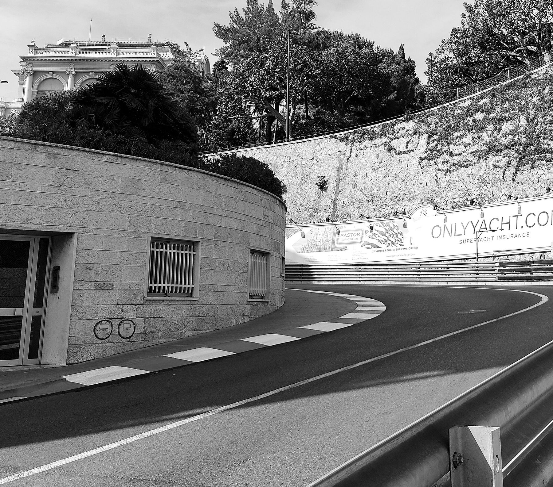Una calle estrecha gira a la derecha entre un edificio de muros de piedra y una barrera de acero, con un piano a rayas en el interior de la curva y una acera que la bordea.