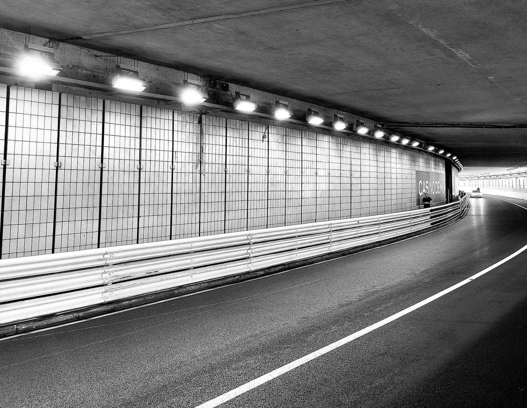 Inside a long, lit road tunnel, a curving carriageway lined with barriers and wire mesh bends away towards daylight at the far end.