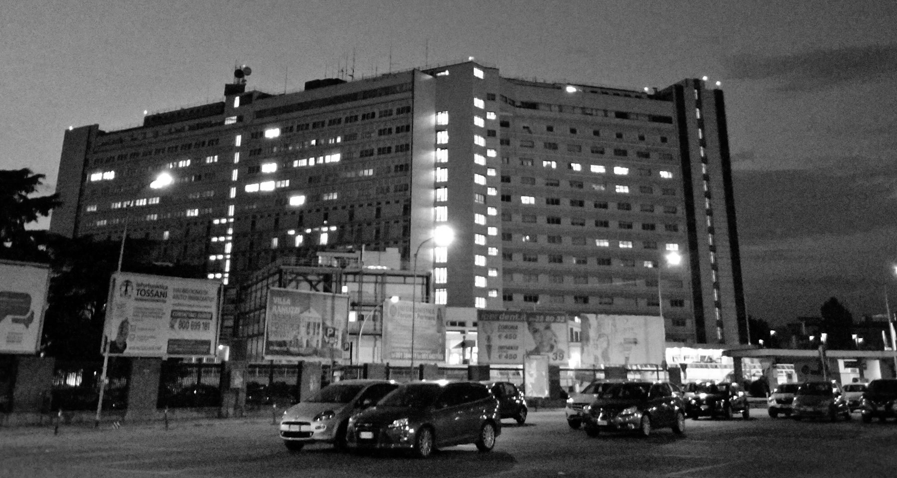 A large hospital block with rows of lit windows stands against an evening sky, billboards and parked cars in front of it.