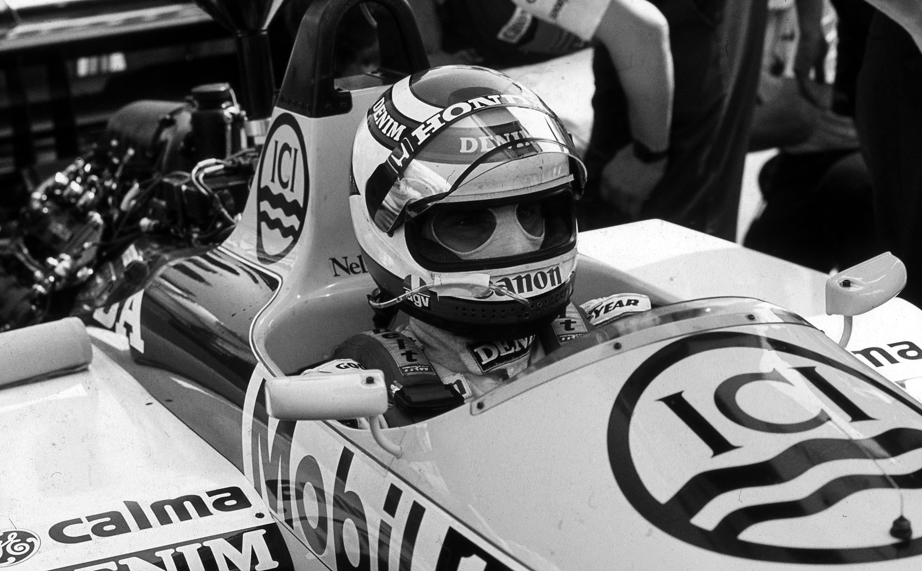 A driver in a helmet with a dark crown and a balaclava sits in the cockpit of a car lettered ‘ICI’, ‘Mobil’ and ‘Canon’ in the pit lane, only his eyes showing, with mechanics around him.