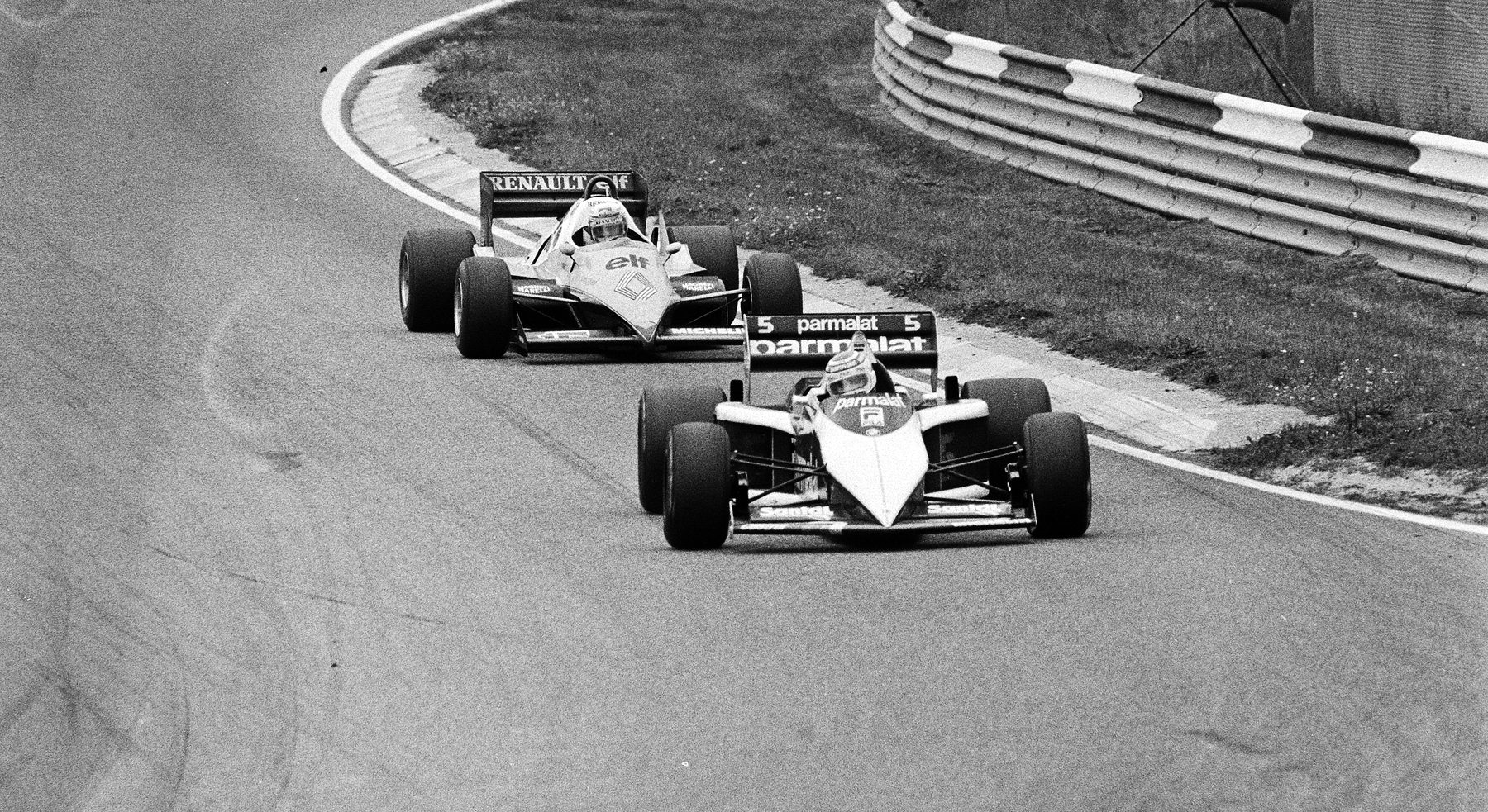 Two Formula One cars sweep through a curve past a triple-rail barrier and a grass verge: a white Brabham, number 5, with a dark rear wing leads a Renault.