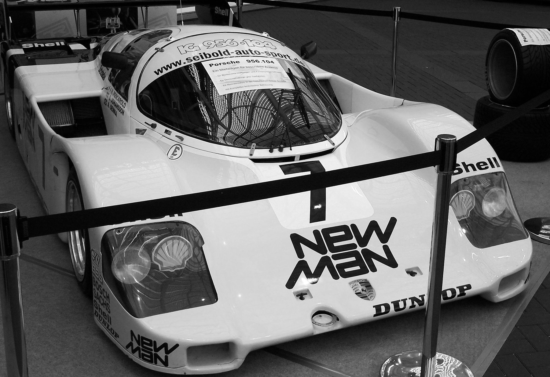 A pale Porsche sports-racing car with a large number 7 and ‘New Man’ lettered on its nose, displayed behind rope barriers at a show.