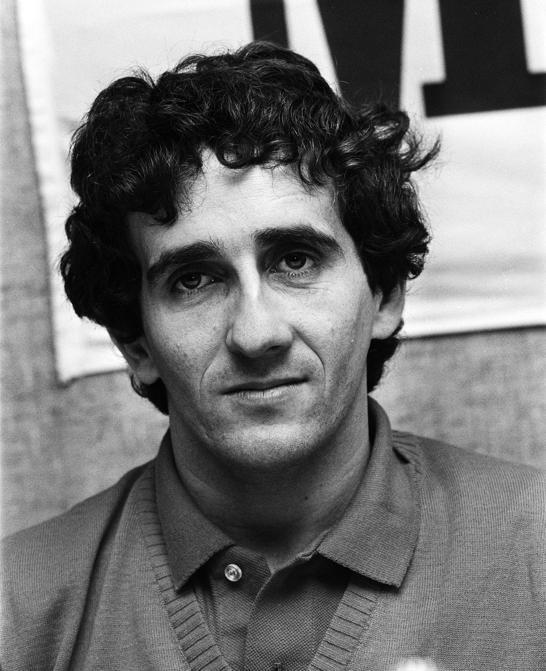 Portrait of a curly-haired man in a polo shirt and V-neck sweater, looking straight at the camera with a faint smile.