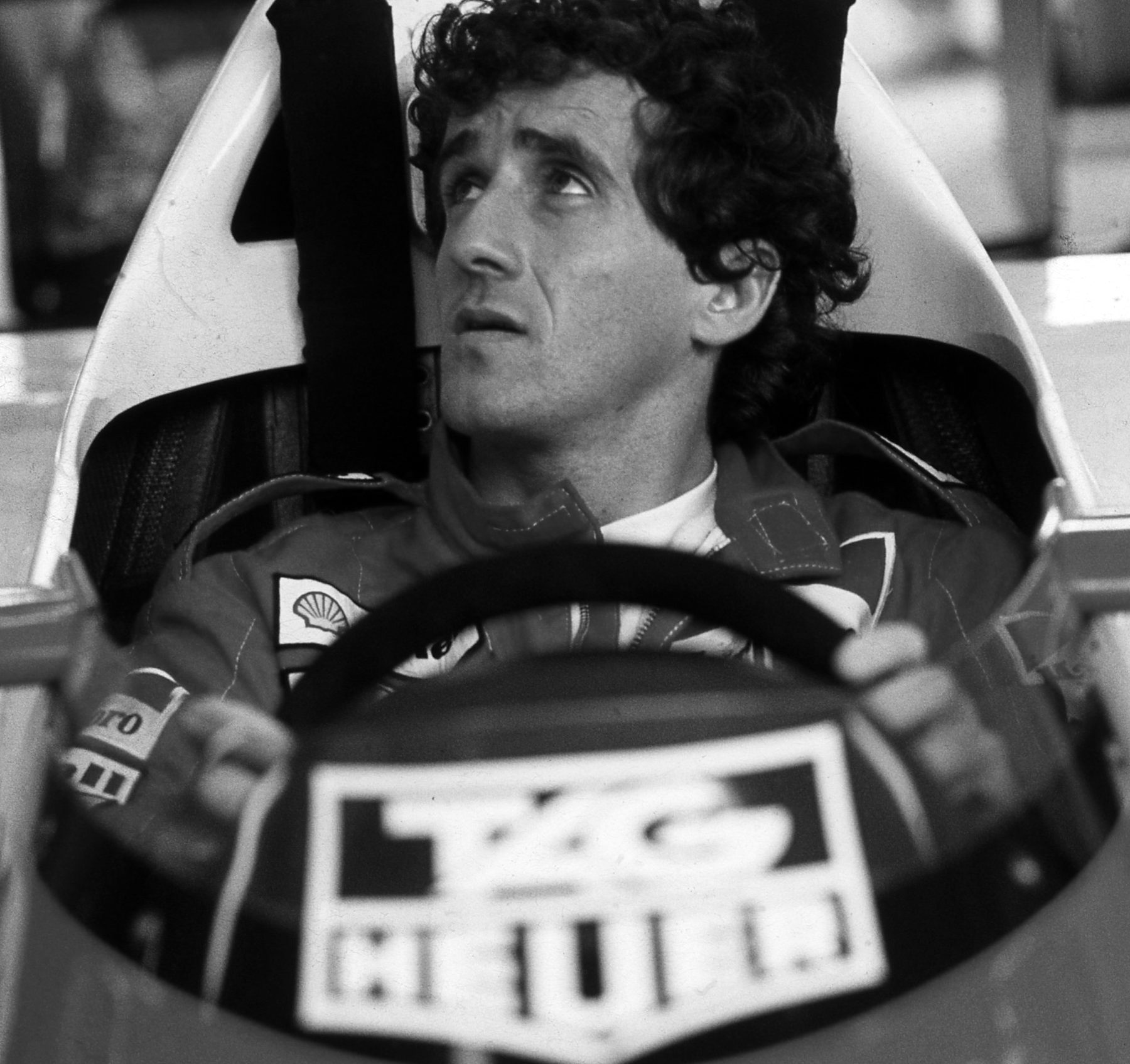 A curly-haired driver sits in a cockpit gripping the steering wheel, looking up and to one side, his helmet not yet on.