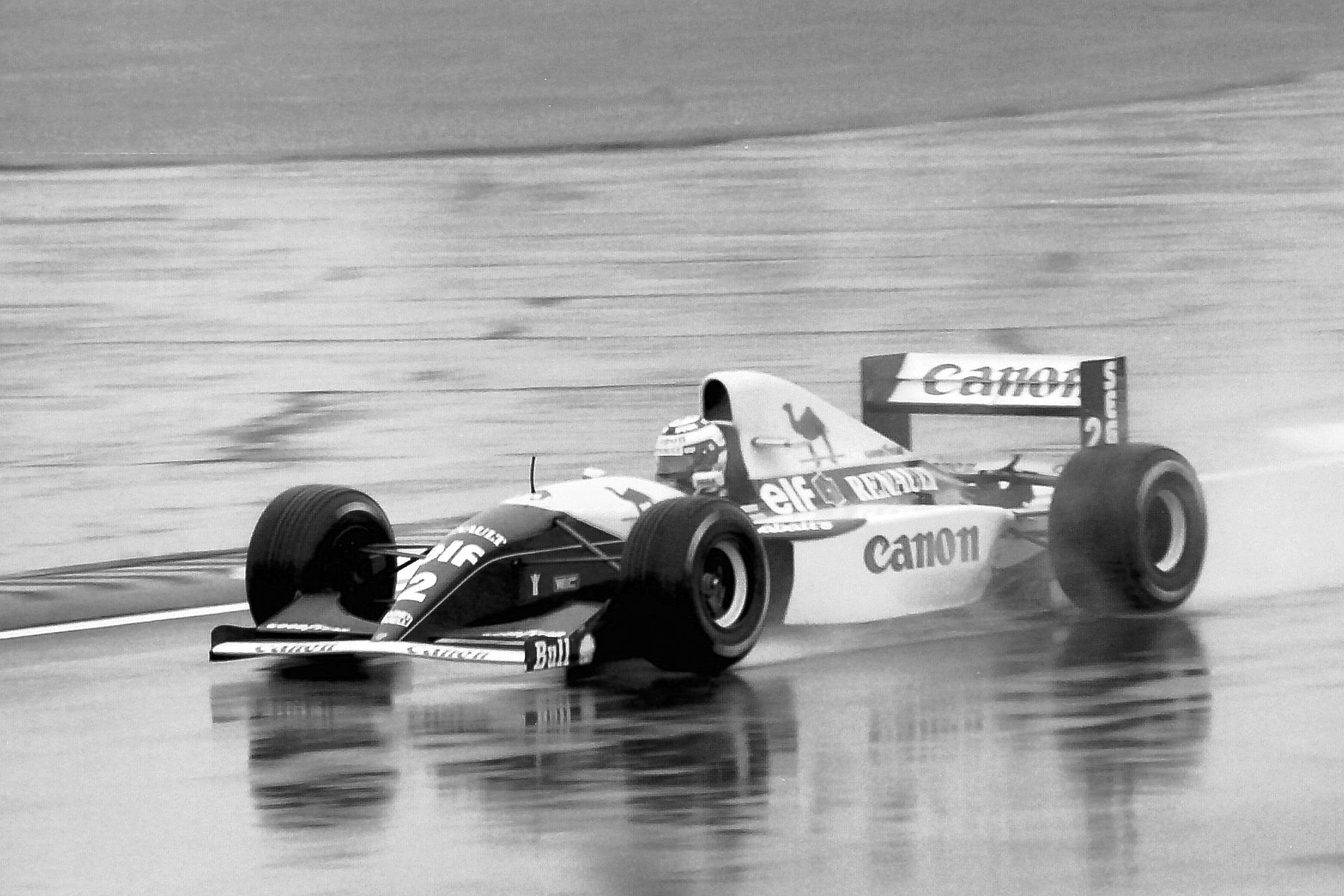 A two-tone Williams, number 2, lettered ‘Canon’ and ‘Renault’, runs through standing water on a wet track, its reflection trailing beneath it.