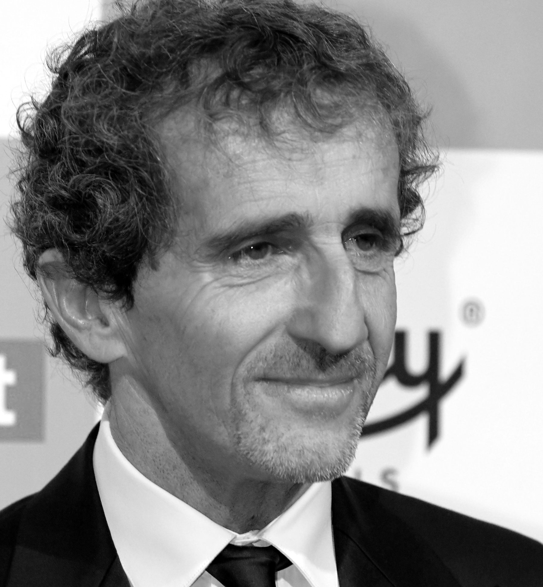 A grey-haired man with curly hair, in a dark suit and tie, smiles slightly as he looks away to the side, in front of a printed backdrop.
