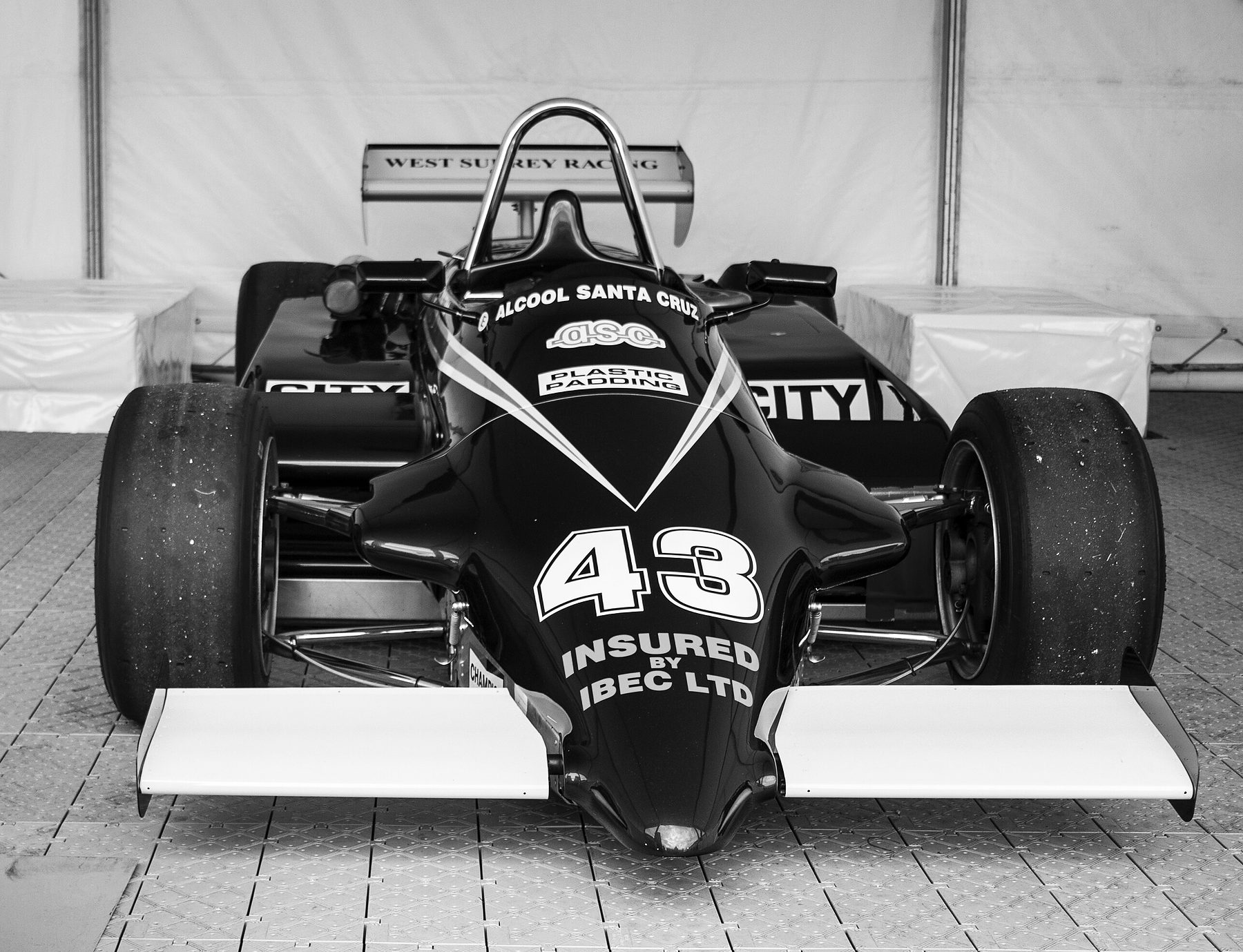 Head-on view of a dark Formula 3 car with a pale front wing and the number 43 on its nose, a roll hoop and rear wing behind the cockpit, parked in a white display tent.