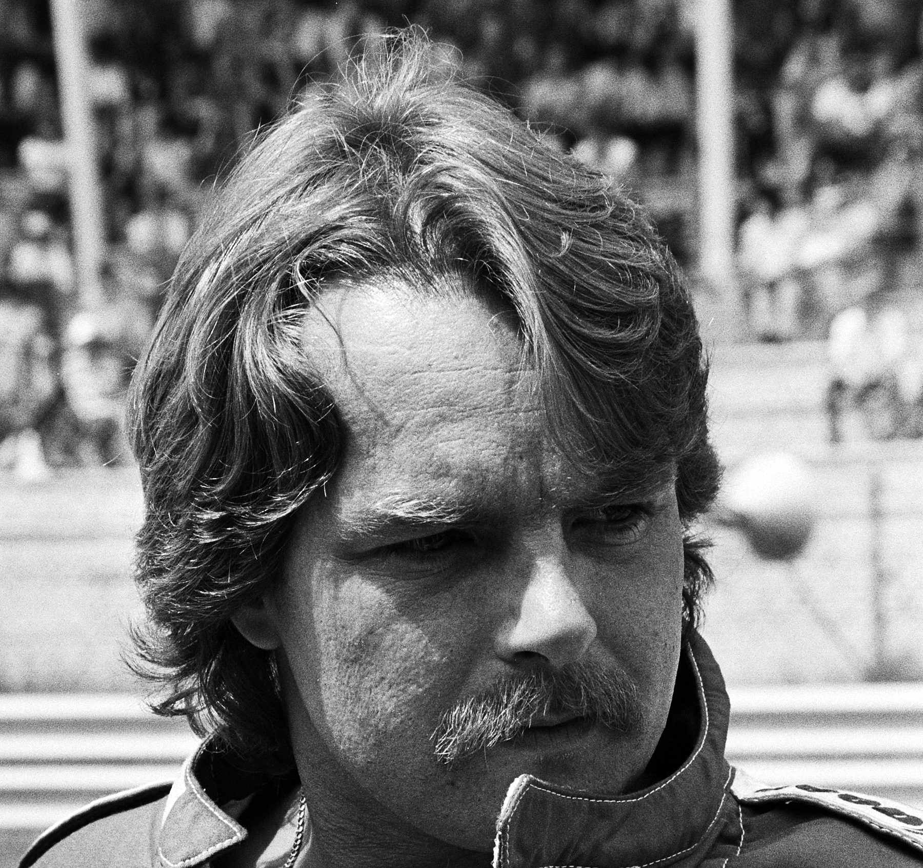 Portrait of a driver with shoulder-length hair and a moustache, collar of his overalls turned up, looking down with a frown.