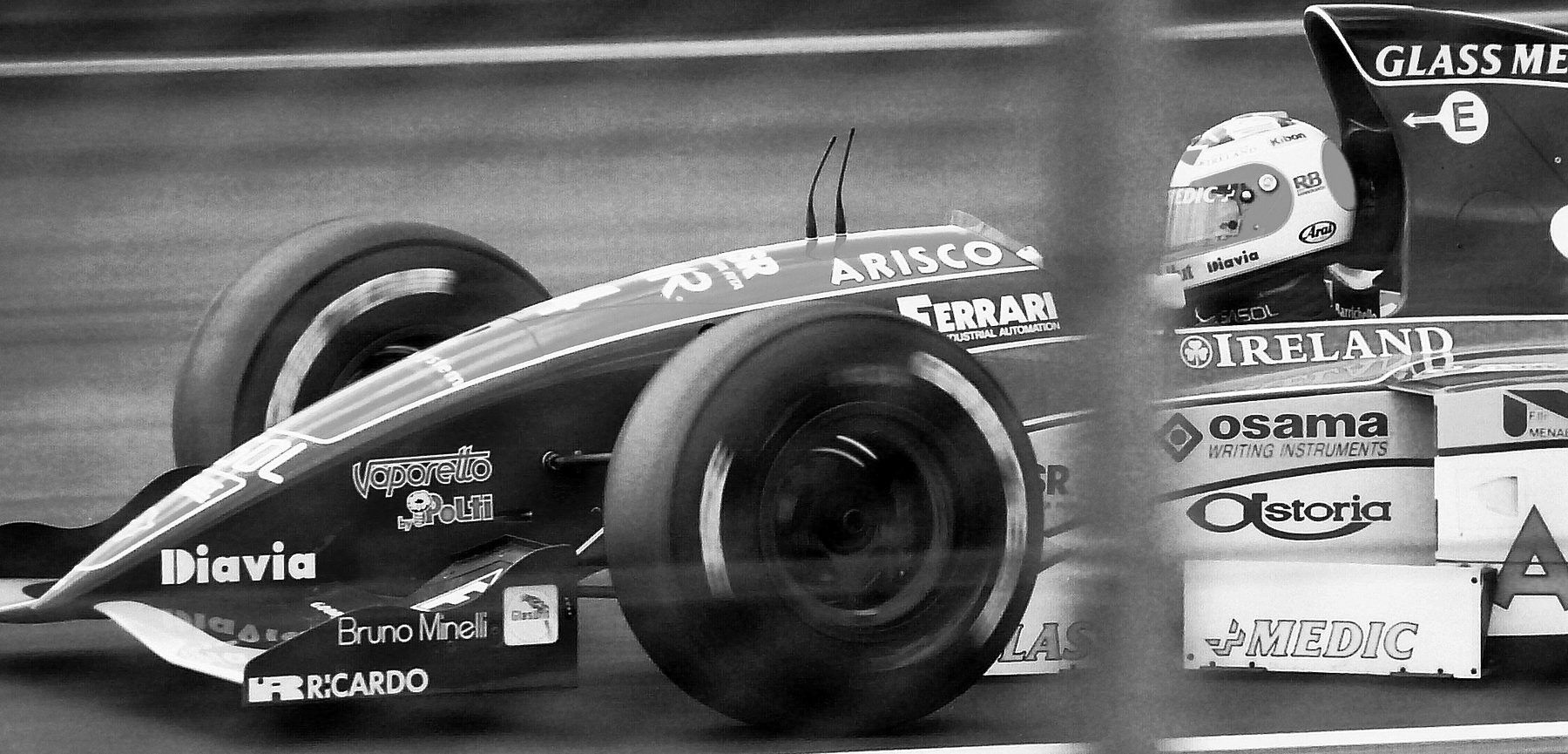 A Jordan Formula One car covered in sponsor lettering passes the camera, the driver’s pale helmet just visible above the cockpit, a blurred post in the foreground.