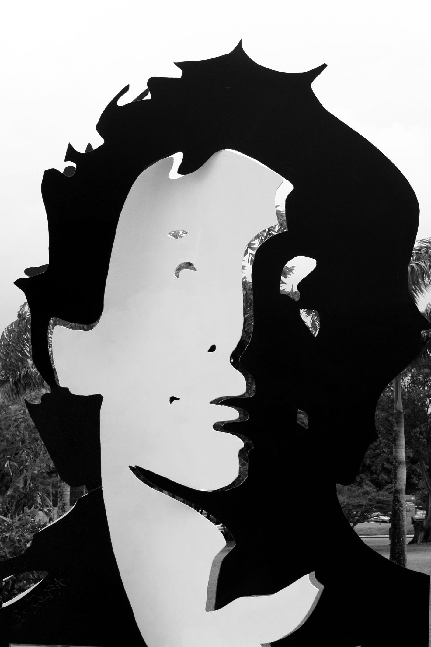 A tall cut-steel portrait of Senna in flat black and white planes, his face half in shadow beneath a jagged mass of hair, with sky and palm trees showing through the cut-outs and the artist’s signature at lower left.