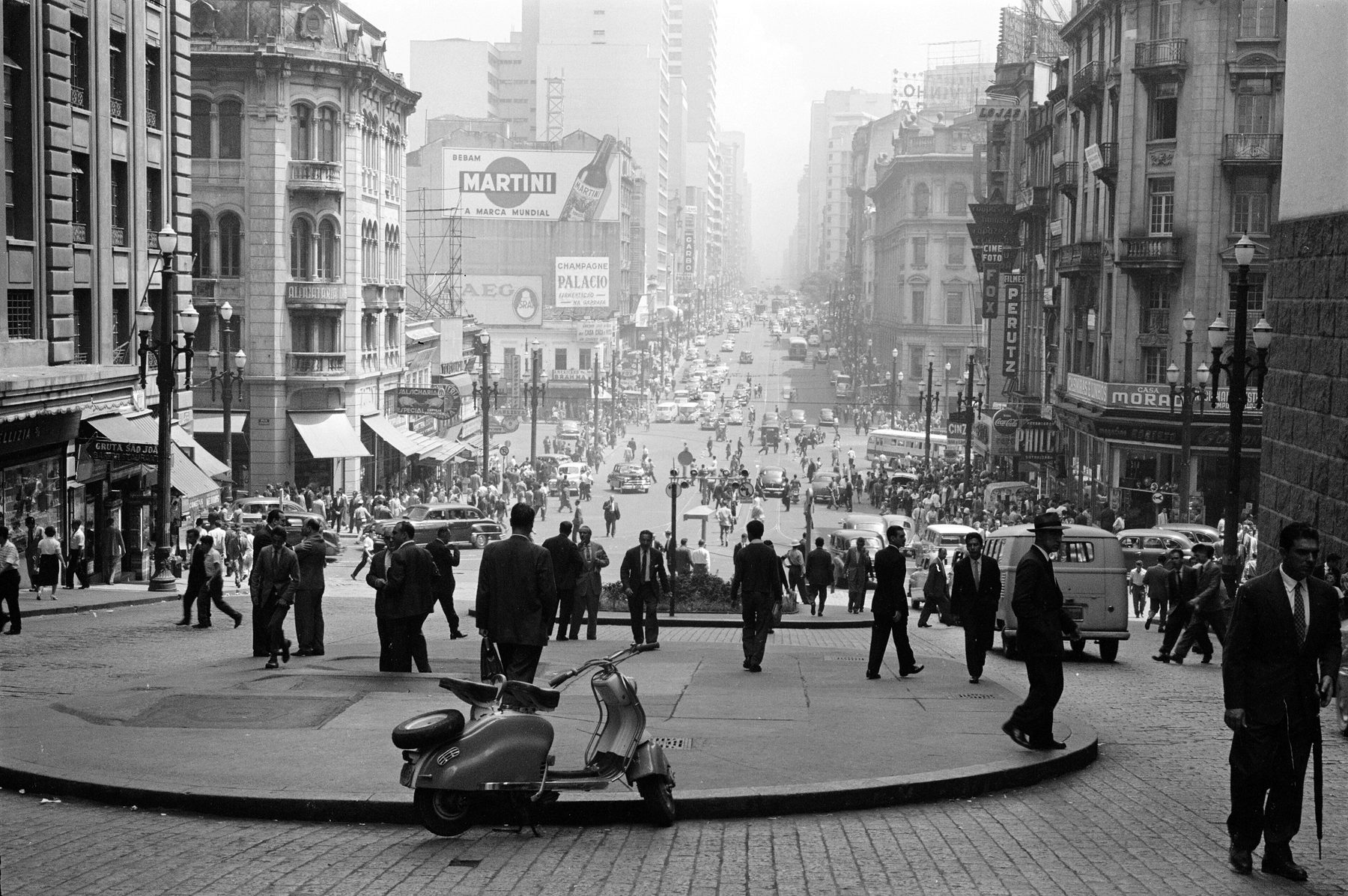 Street scene: men in suits and hats walk across a paved square at the foot of a steep city street lined with tall buildings and signs, a motor scooter parked in the foreground.