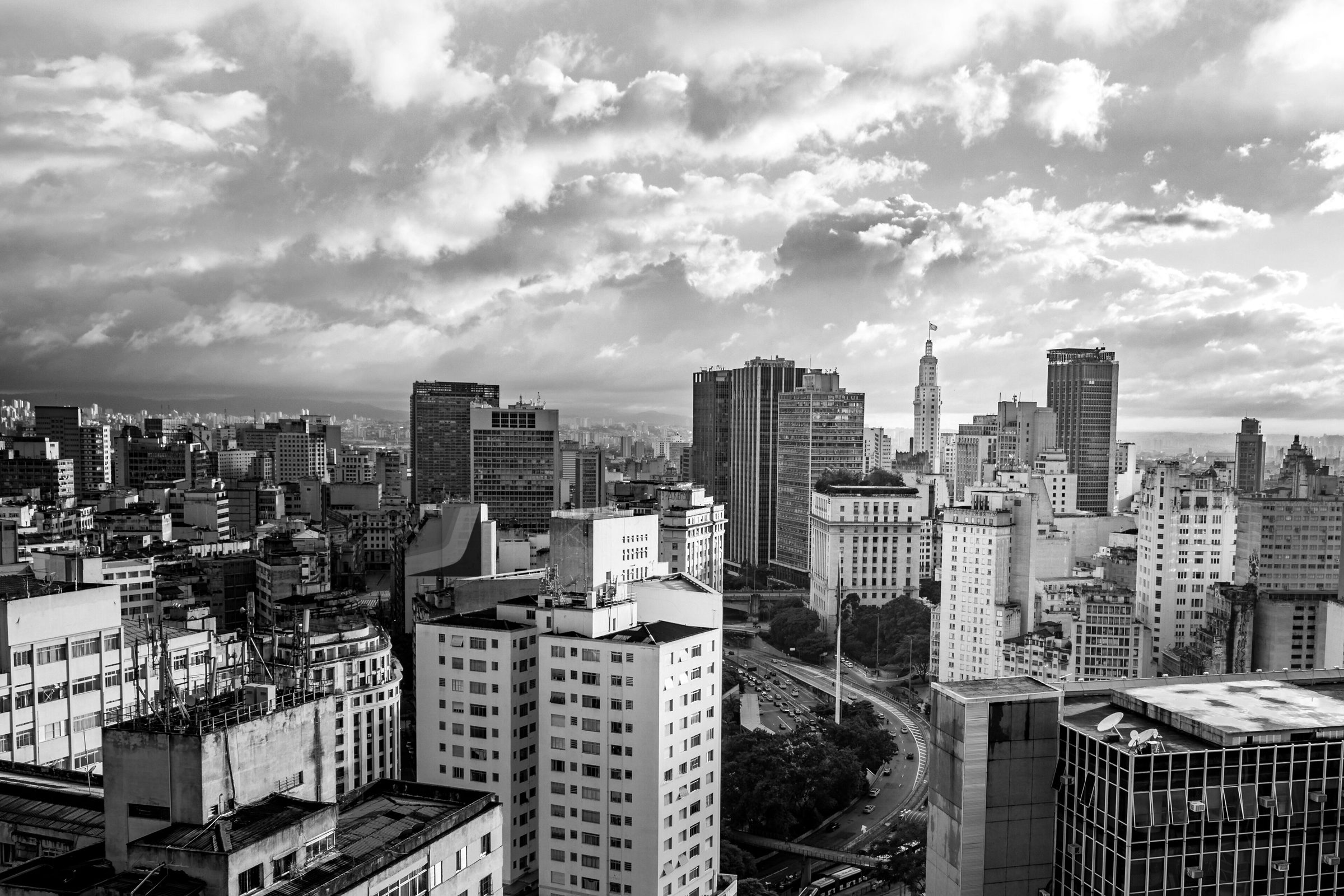 The towers of a dense city centre under a sky of towering, sunlit clouds, seen from a high rooftop.
