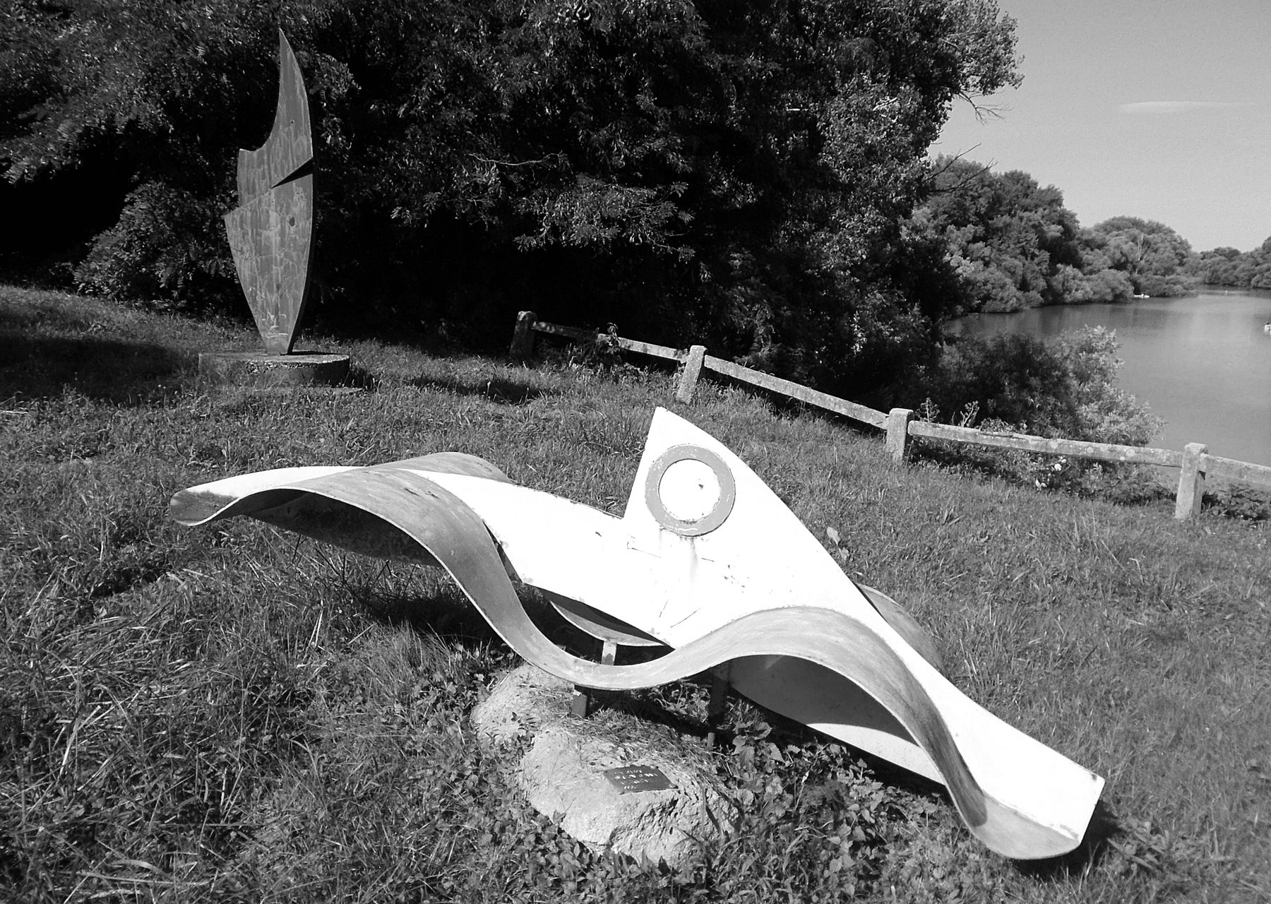 A low, pale sculpture of painted iron, a pointed wedge with a ring wrapped by a wavy steel ribbon, rests on a boulder on a grassy riverbank; a tall dark metal form stands behind it and a river runs beyond the trees.