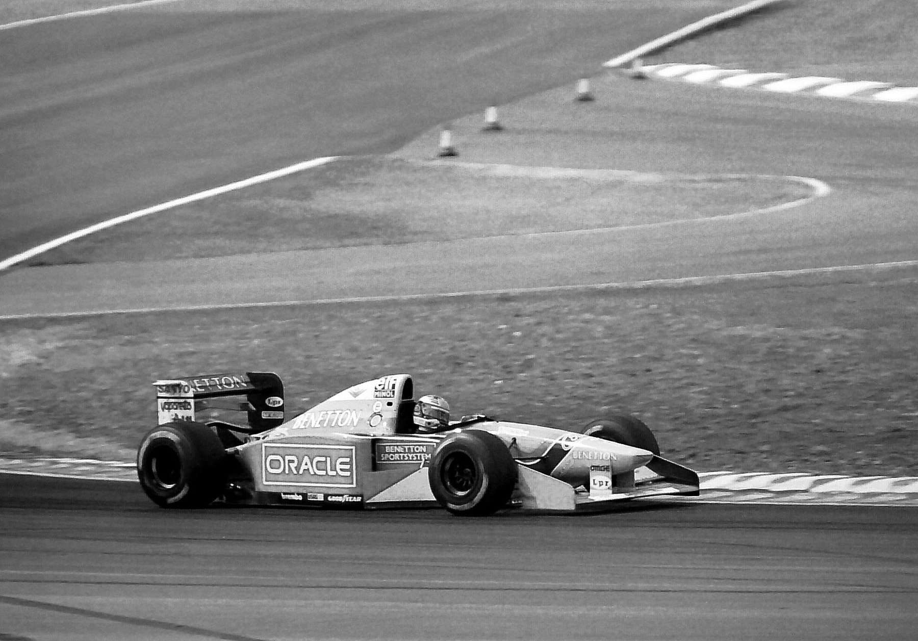 A pale Benetton Formula One car with dark lettering on its sidepods sweeps round a bend on a wide circuit, grass and kerbs around it and open tarmac behind.