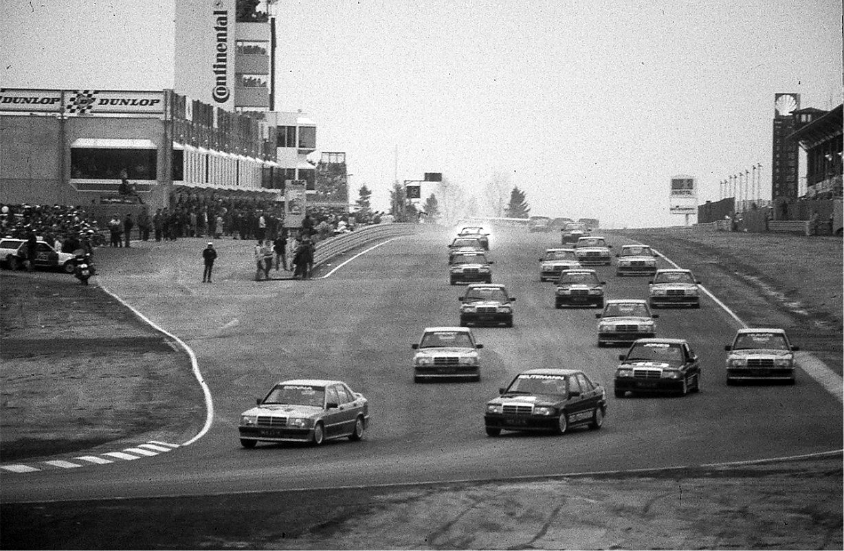 A pack of identical Mercedes saloons streams down a wide straight towards the camera past a grandstand and pit building on a dull day.