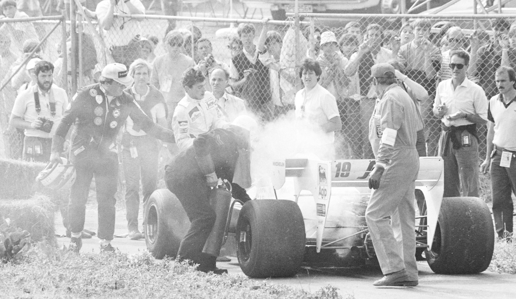 A young dark-haired driver in white overalls, his helmet off, stands beside his smoking white car as a marshal bends over it, a crowd pressed against the wire fence behind.