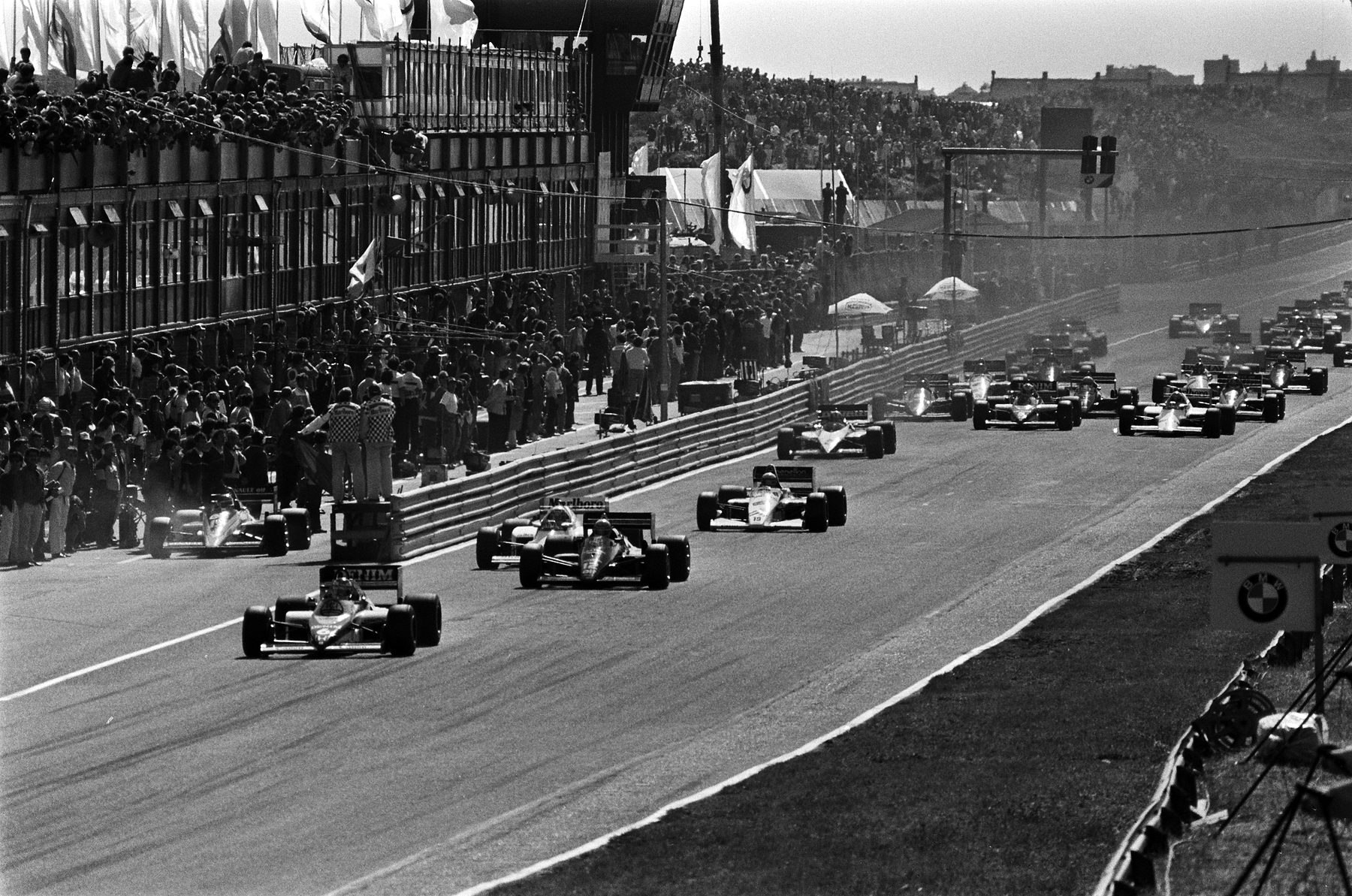 The start of a Grand Prix seen from the side, a pack of cars pouring down the long pit straight past packed grandstands and the pits.
