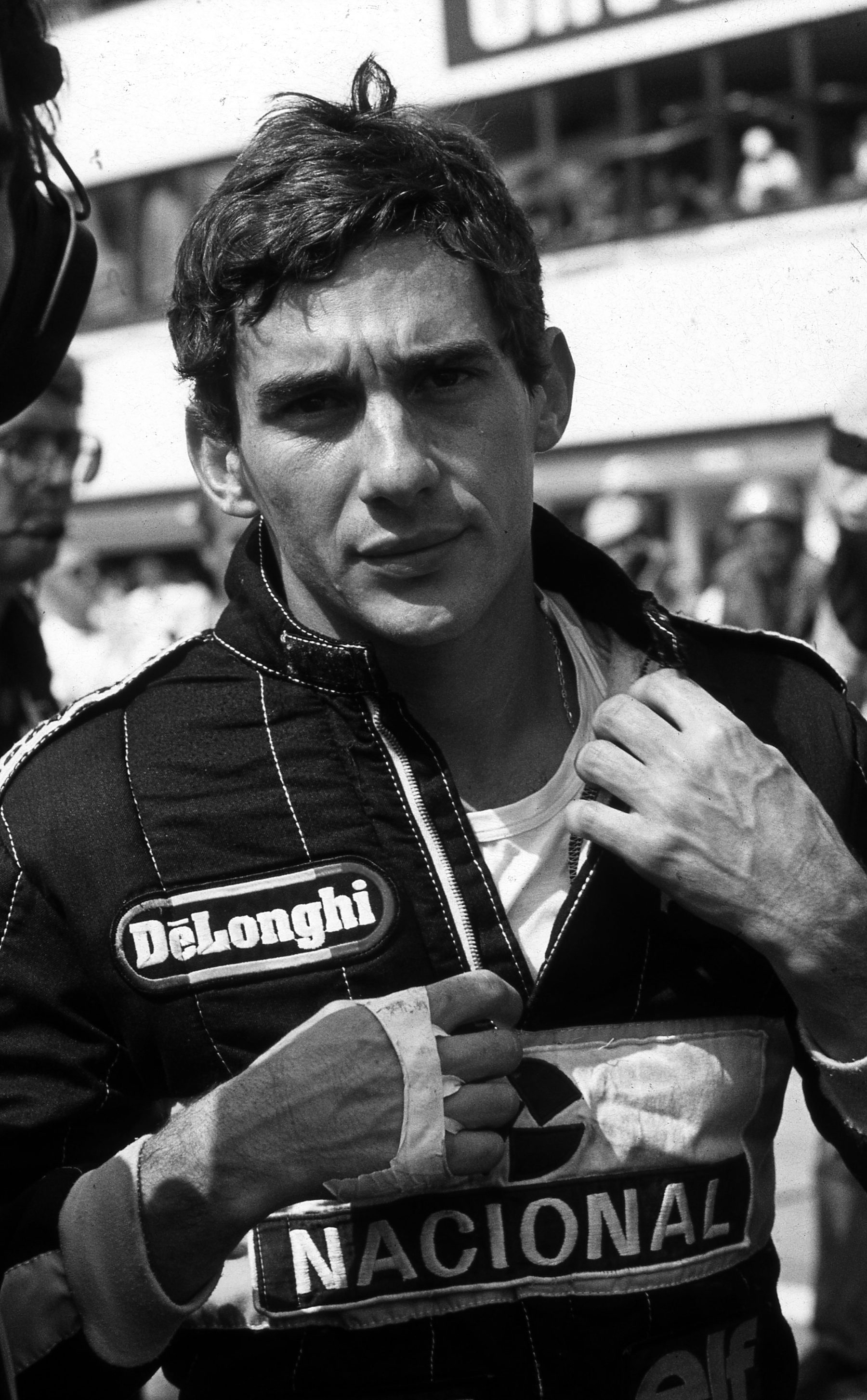 A dark-haired young man in black racing overalls with sponsor patches looks straight into the lens, one hand at his collar, the crowded pit lane blurred behind him.