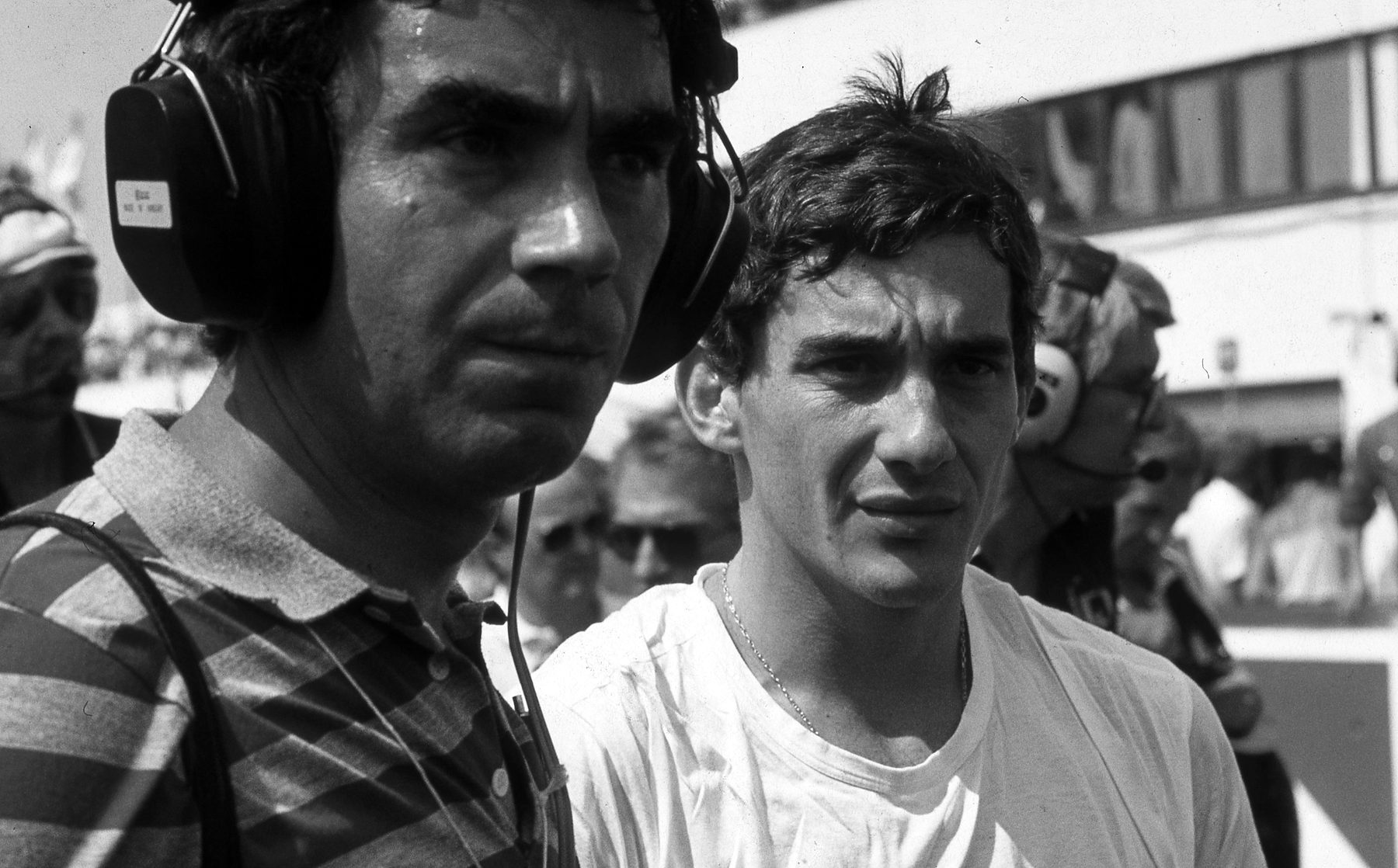 Two men side by side in a busy pit lane: a man in a striped shirt wearing headphones, and a young dark-haired man in a plain white T-shirt looking past the camera.