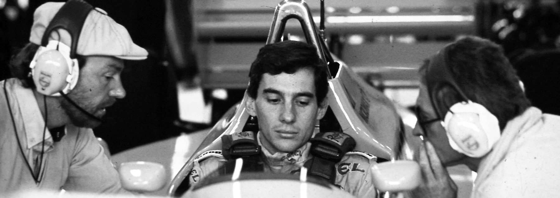 A young driver sits in the cockpit of his car in the garage, eyes lowered, while two engineers in headphones lean in from either side, one of them speaking.