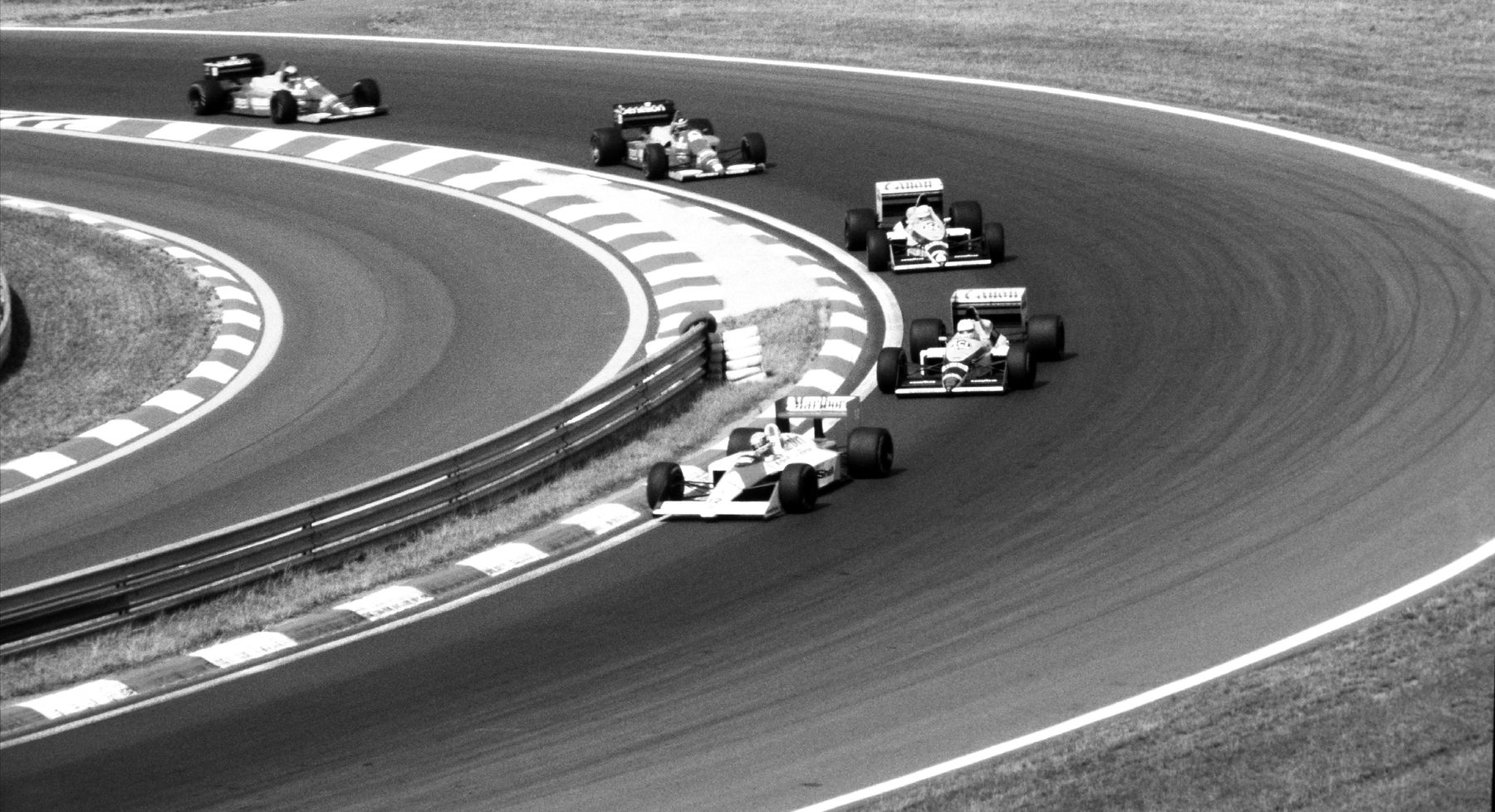 Seen from a high bank, a pale McLaren leads a line of cars round a long hairpin edged with striped kerbs.
