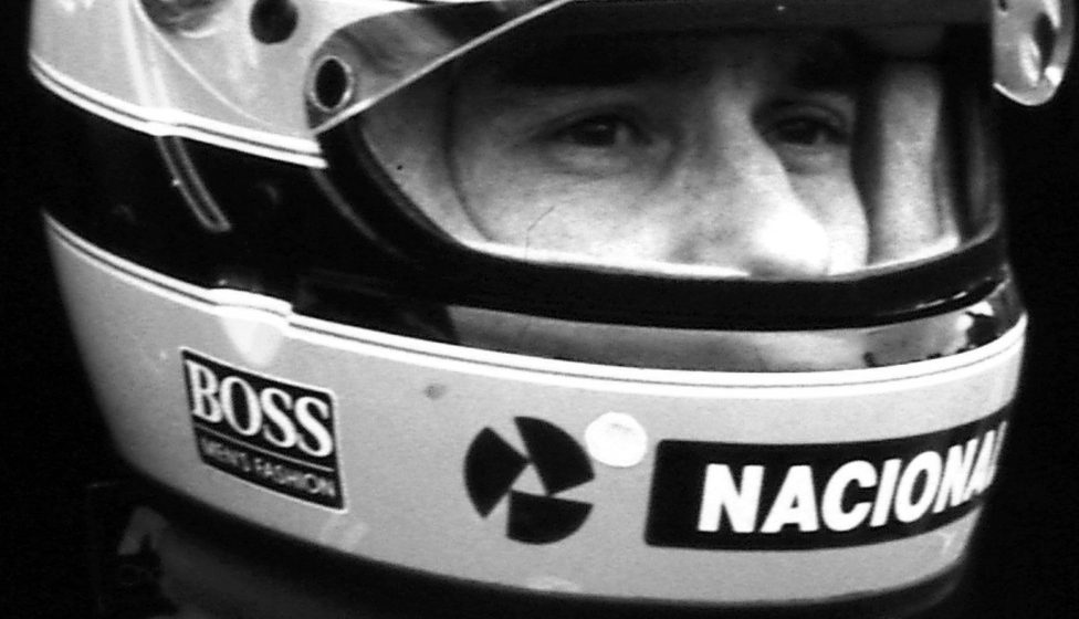 A driver’s eyes look out from the opening of his striped helmet, the rim of the raised visor above them, ‘Boss’ and ‘Nacional’ on the chin guard.