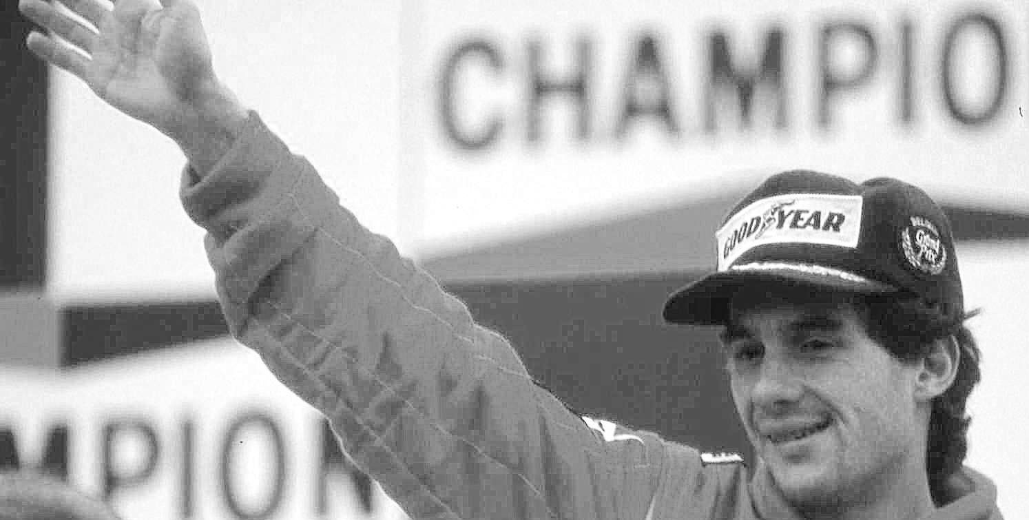A smiling driver in a sponsor’s cap raises his arm high in a wave from a podium, a large ‘Champion’ backdrop behind him.
