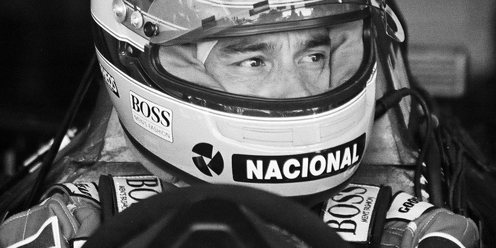 A driver’s eyes glance to one side through the open visor of his helmet, ‘Nacional’ across its chin guard and ‘Boss’ on its side and on his shoulders, the steering wheel dark in the foreground.