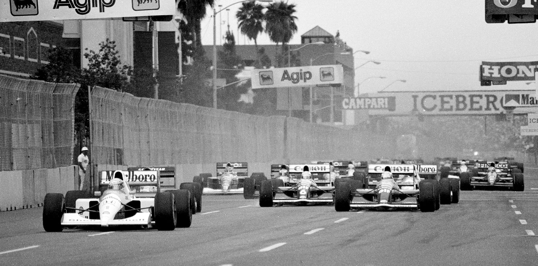 A field of Formula One cars streams away from the start down a straight between concrete walls and hoardings, palm trees and buildings behind.