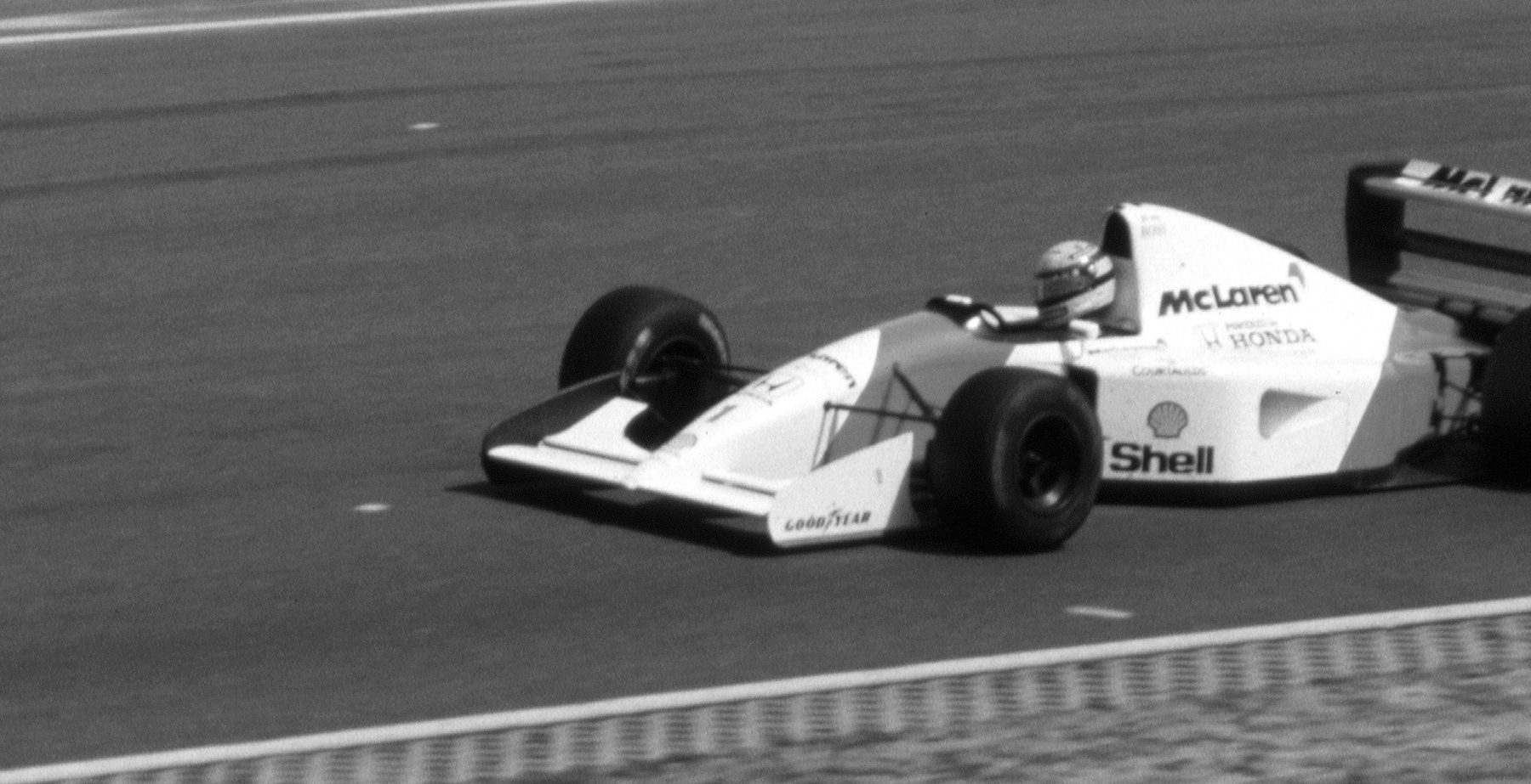 A pale McLaren runs along a wide, empty stretch of track, the driver’s helmet just above the cockpit, kerbs along its edge and the rear of another car at the right edge.
