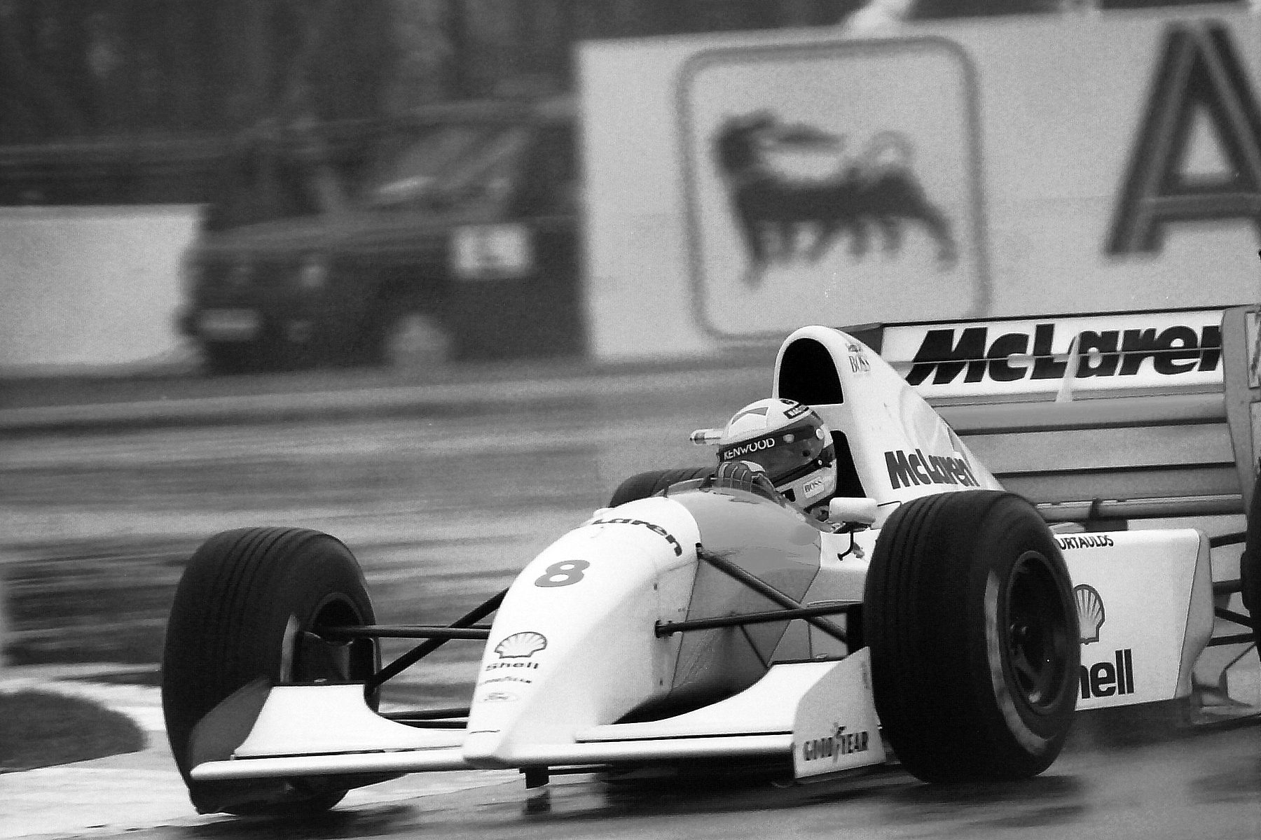 A pale McLaren numbered 8 corners on a wet track, the driver’s striped helmet sharp in the cockpit, a blurred hoarding with a large emblem behind.