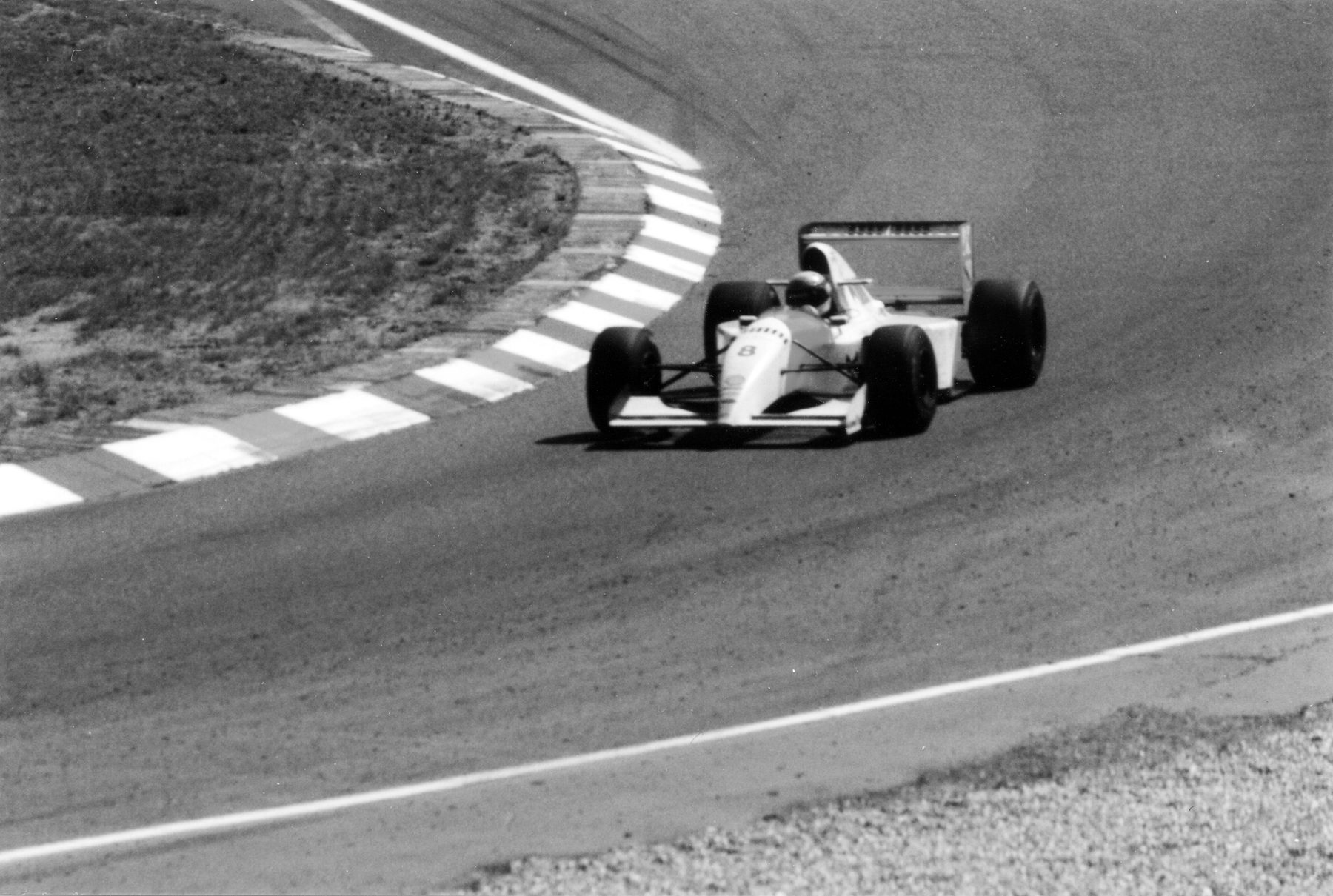 Seen from above, a pale McLaren swings out of a long curve bordered by striped kerbs on an empty, sunlit circuit.