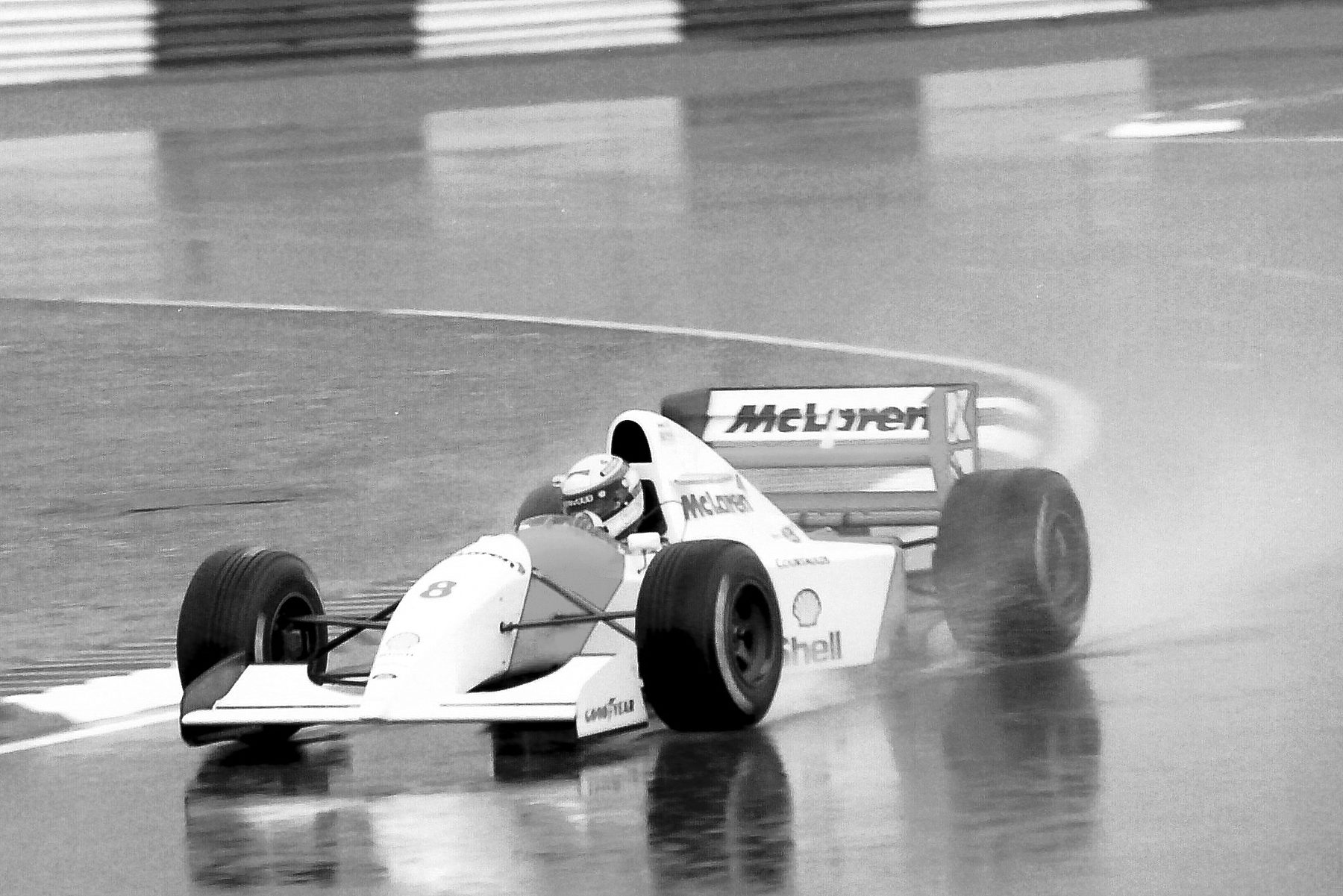 A pale McLaren sweeps through a bend on a streaming wet track, throwing up spray, its reflection shimmering in the water beneath it.