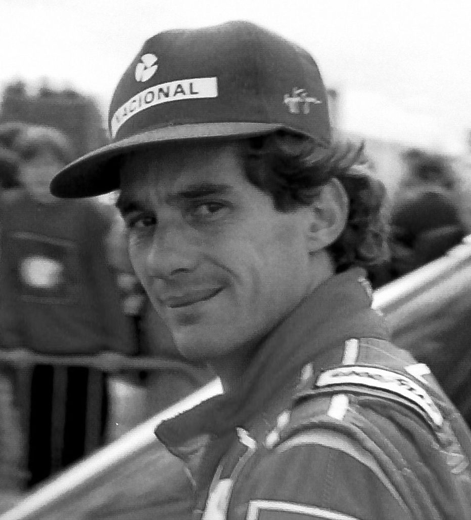 A man in a ‘Nacional’ cap and racing overalls glances back over his shoulder at the camera with a small, knowing smile, spectators blurred behind a fence.